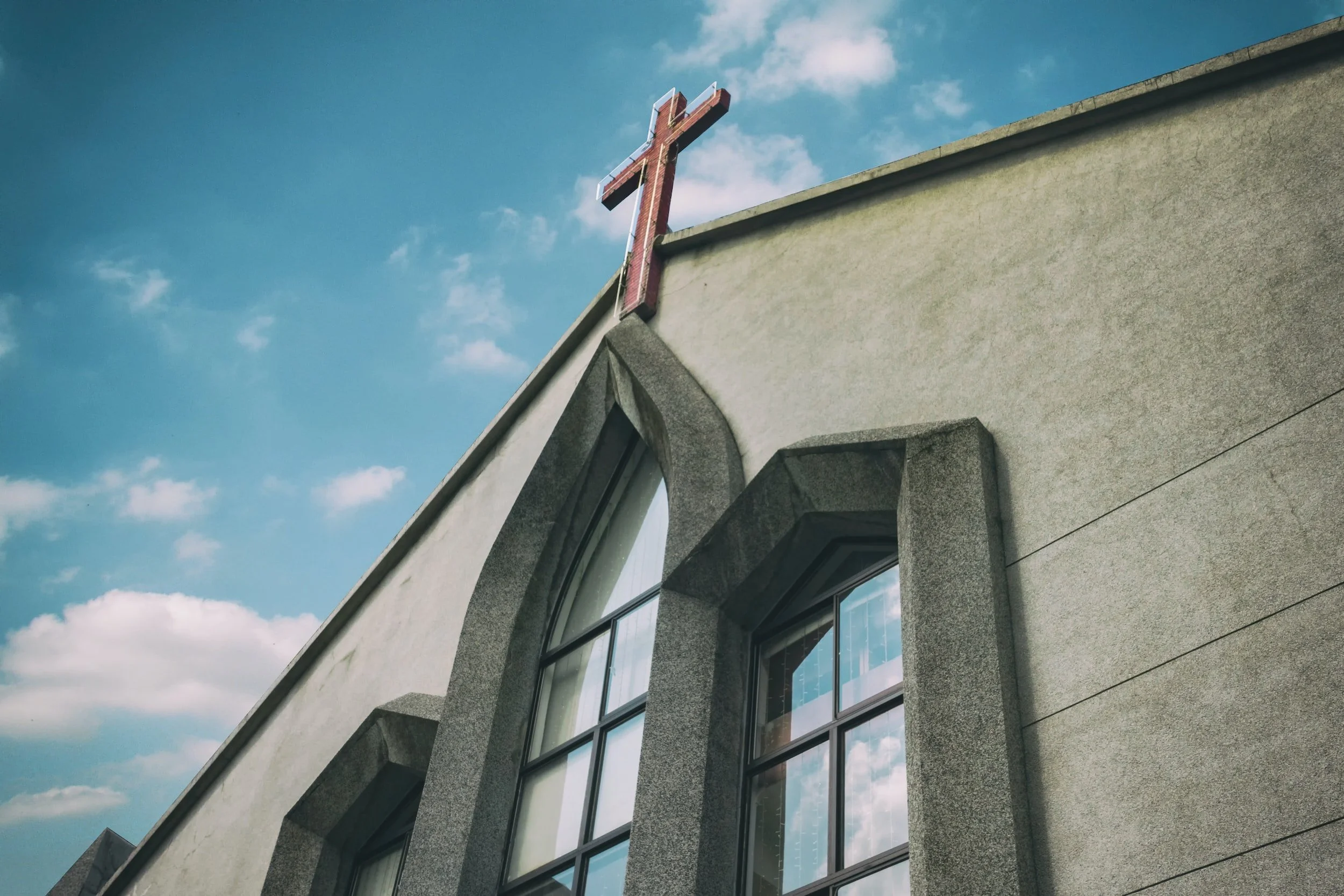 Close-up of church building with stained glass windows and a red cross on top, against a blue sky with clouds.