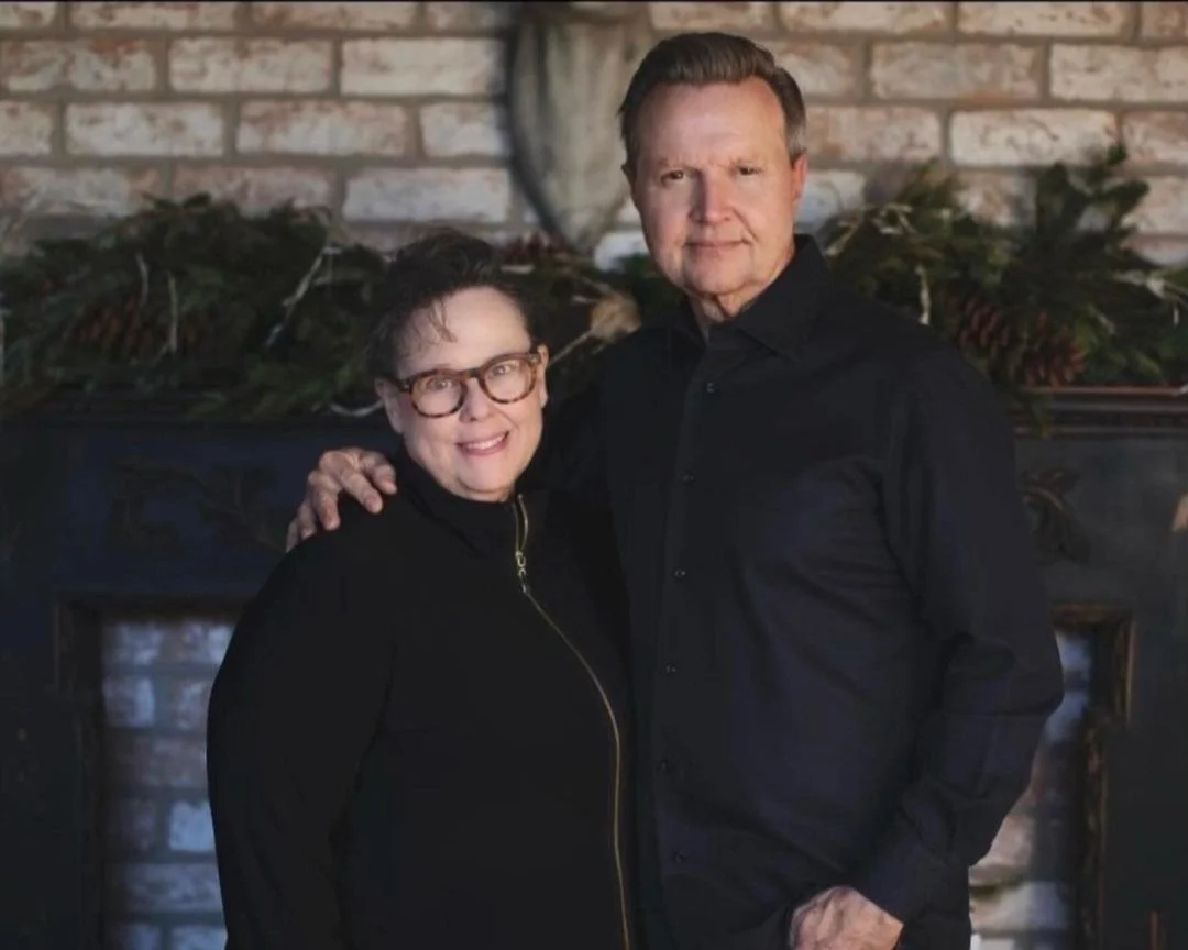 A woman with glasses and a man in black shirts standing in front of a brick wall with holiday greenery and pinecones.