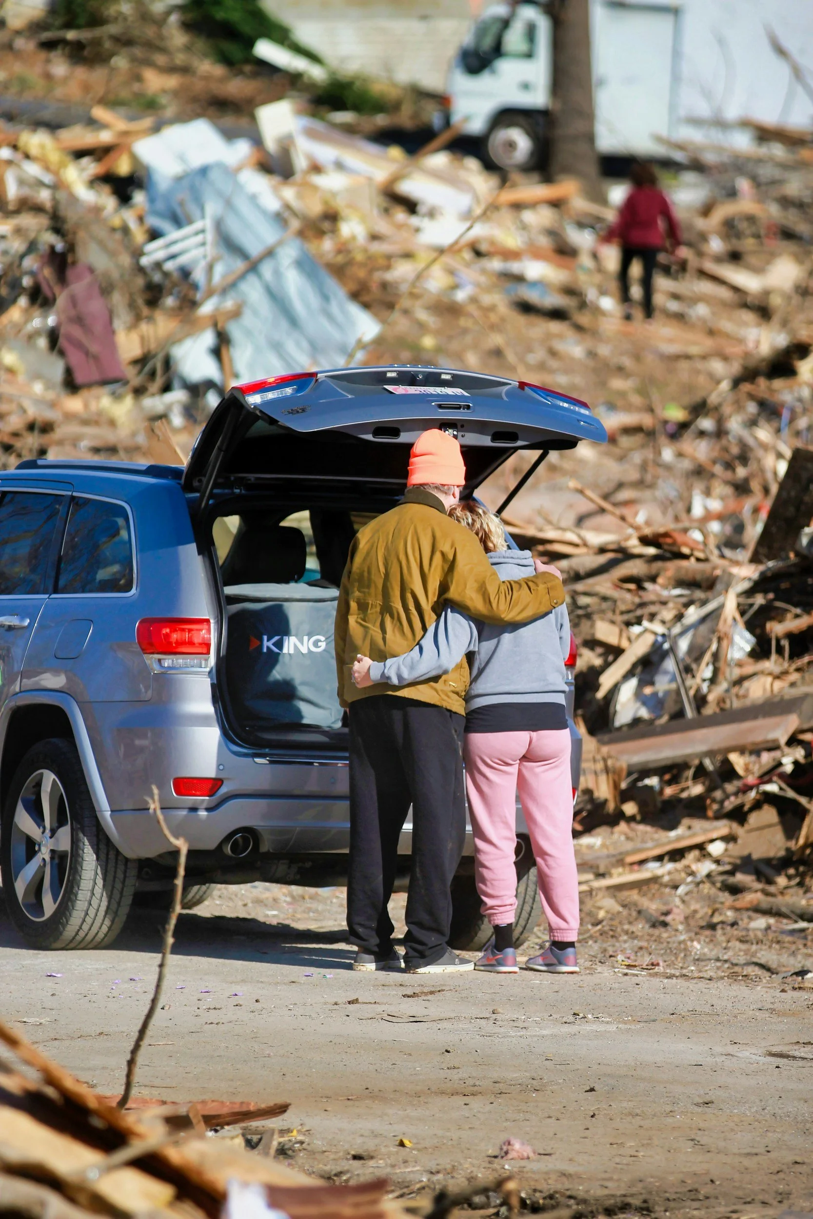 A couple embraces by their gray SUV with the trunk open, in front of a large debris field from a natural disaster with damaged buildings and downed trees, and a person walking among the wreckage in the background.