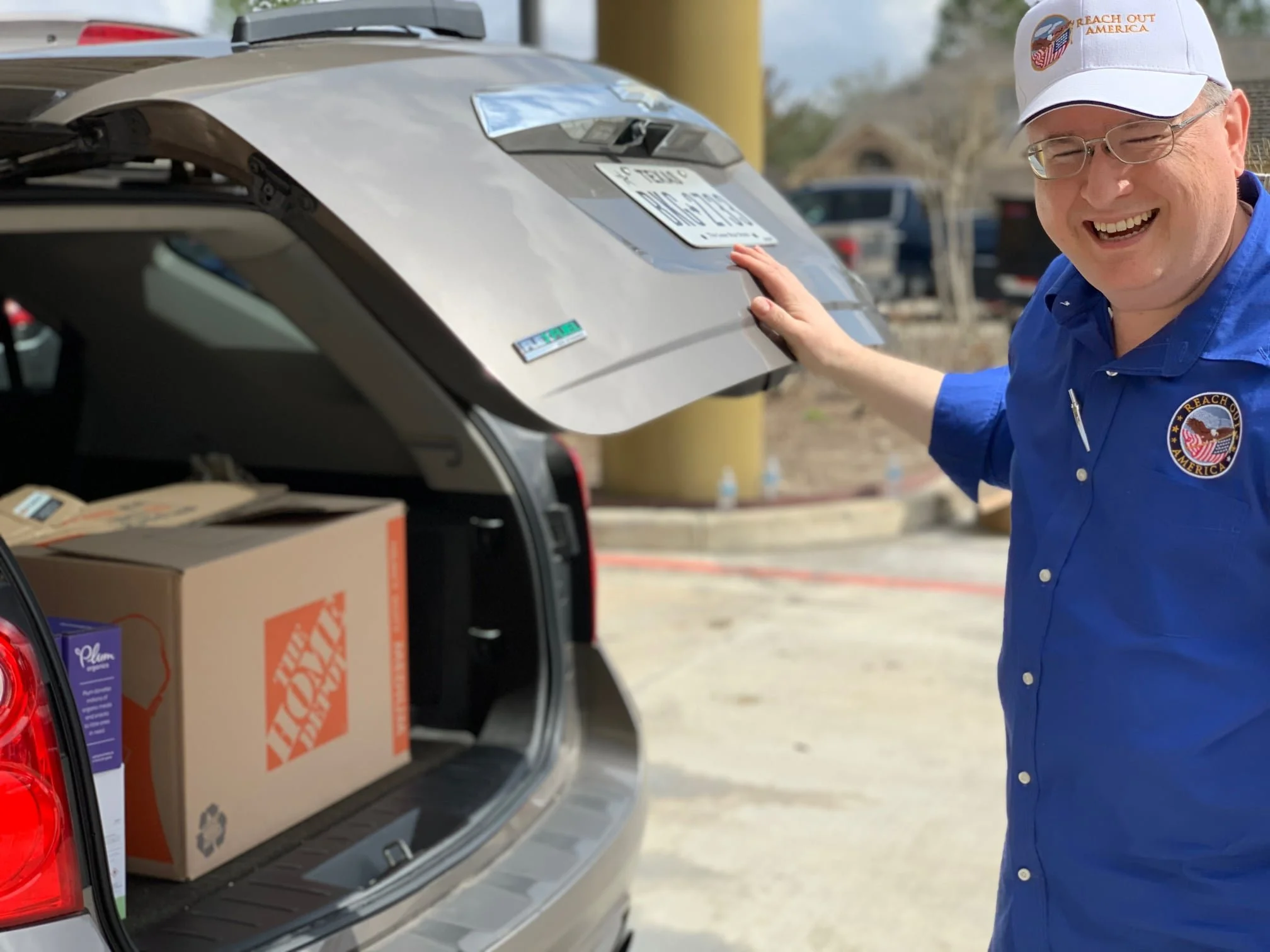 A man with glasses wearing a blue uniform and a white cap with an America logo is smiling and opening the trunk of a car filled with boxes including a Home Depot box.