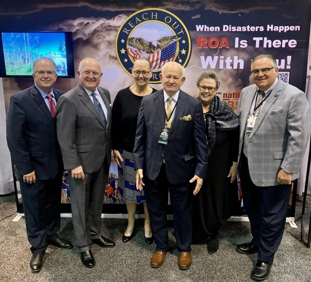 Group of six professionally dressed people standing in front of a display with a logo reading "Reach Out" and a sign that says "When Disasters Happen ROA Is There With You!" at an event.