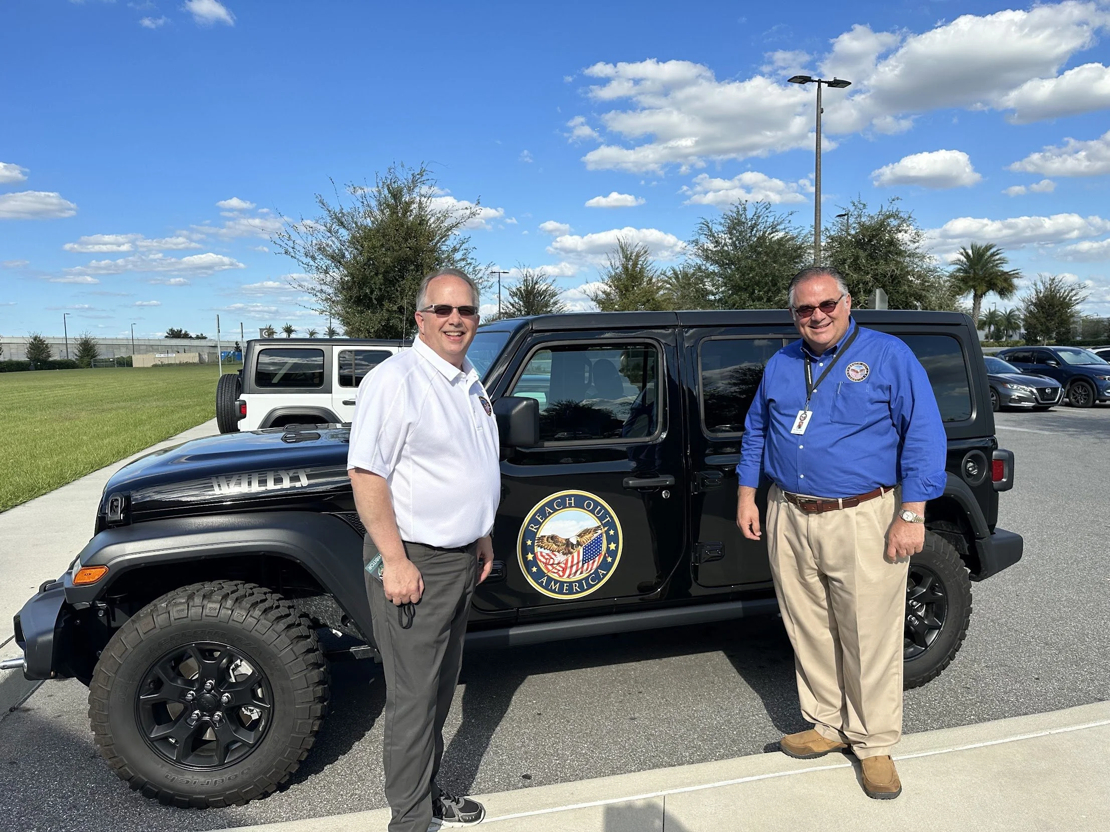 Two men standing beside a black Jeep with a 'Reach Out America' logo, in an outdoor parking lot with trees and a blue sky with clouds in the background.