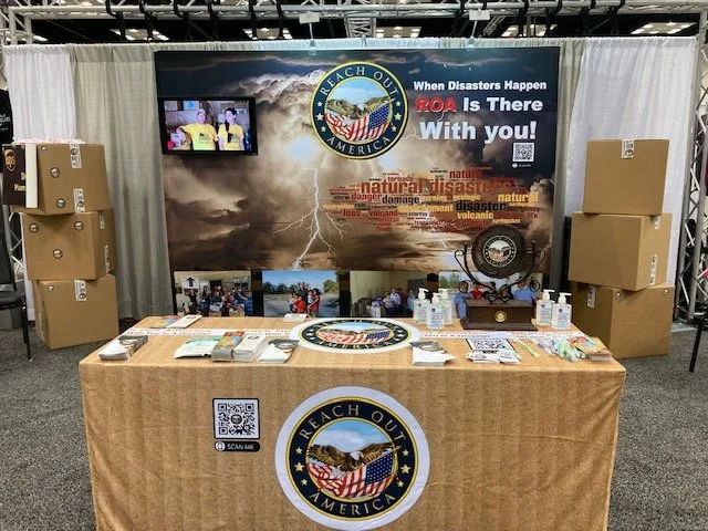 Booth setup for Reach Out America with a table covered in literature and branded items, backdrop featuring storm imagery and a logo, large boxes on either side, and promotional materials on the table.