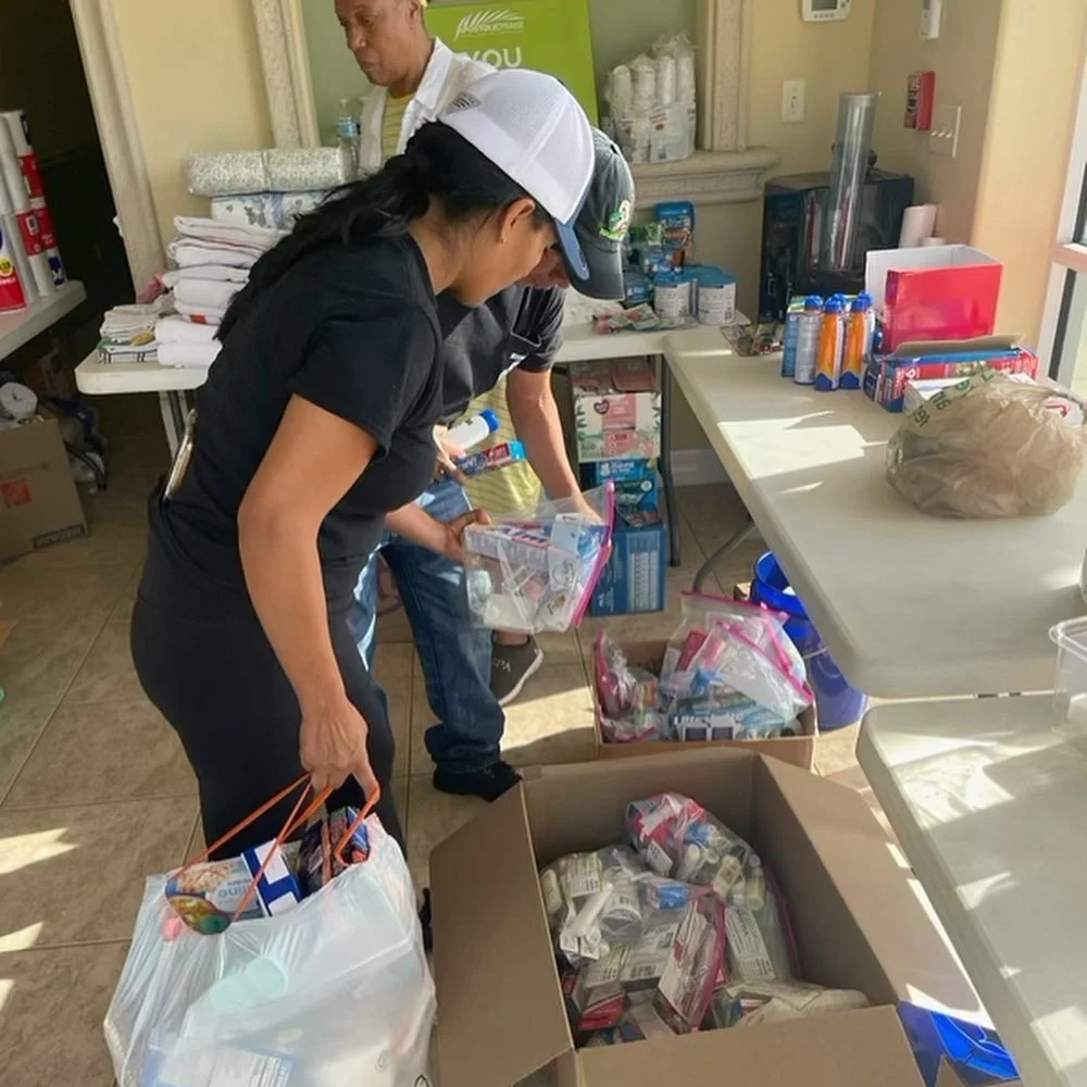 People sorting food supplies and packaged items in a room, with boxes and bags of groceries, canned goods, and snacks on tables and the floor.