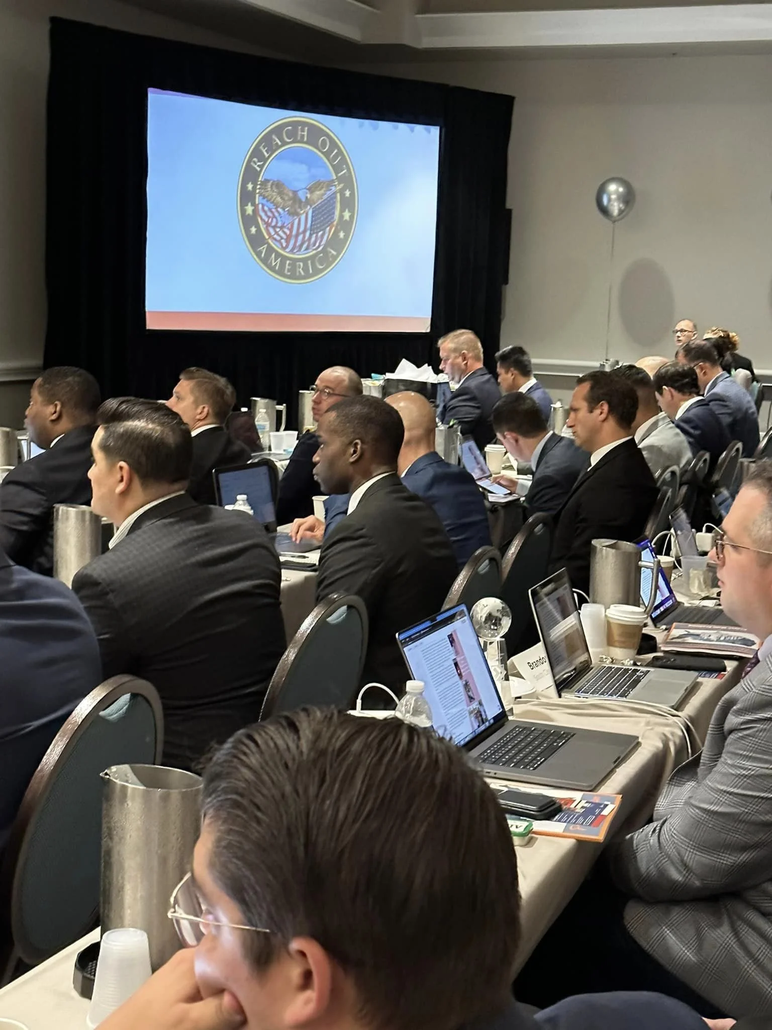Business conference with attendees seated at long tables, looking at laptops, and a large screen displaying the Reach Out America logo, which features a bald eagle holding an American flag in its beak, with a blue sky background.