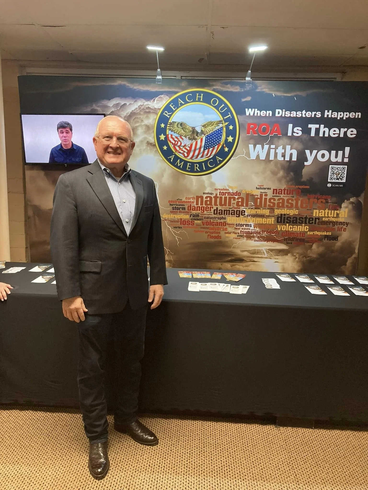 A man in a black suit standing in front of a booth with a backdrop promoting disaster relief for America, featuring a logo with an eagle, American flag, and the words "Reaching Out America." The booth has brochures and pamphlets on a table.
