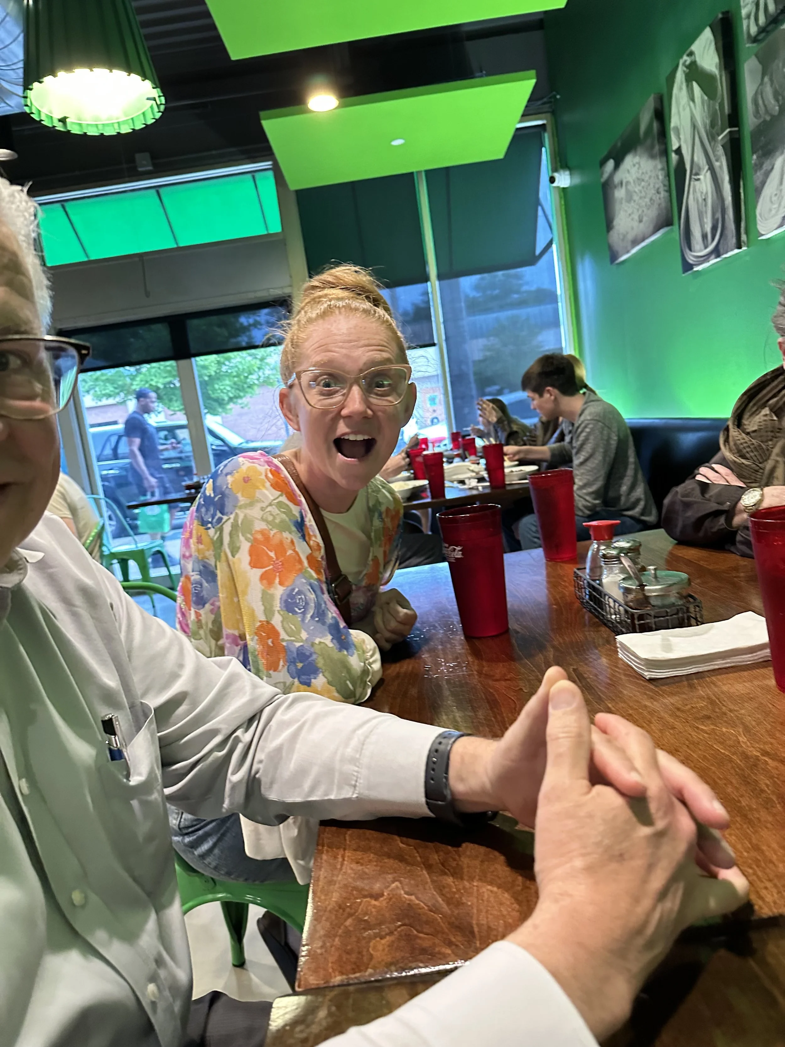 A red-haired woman with glasses and a floral top sitting at a restaurant table, surprised and smiling, with a man partially visible to her left, holding hands with another person. In the background, other diners are seated, and the restaurant has gre