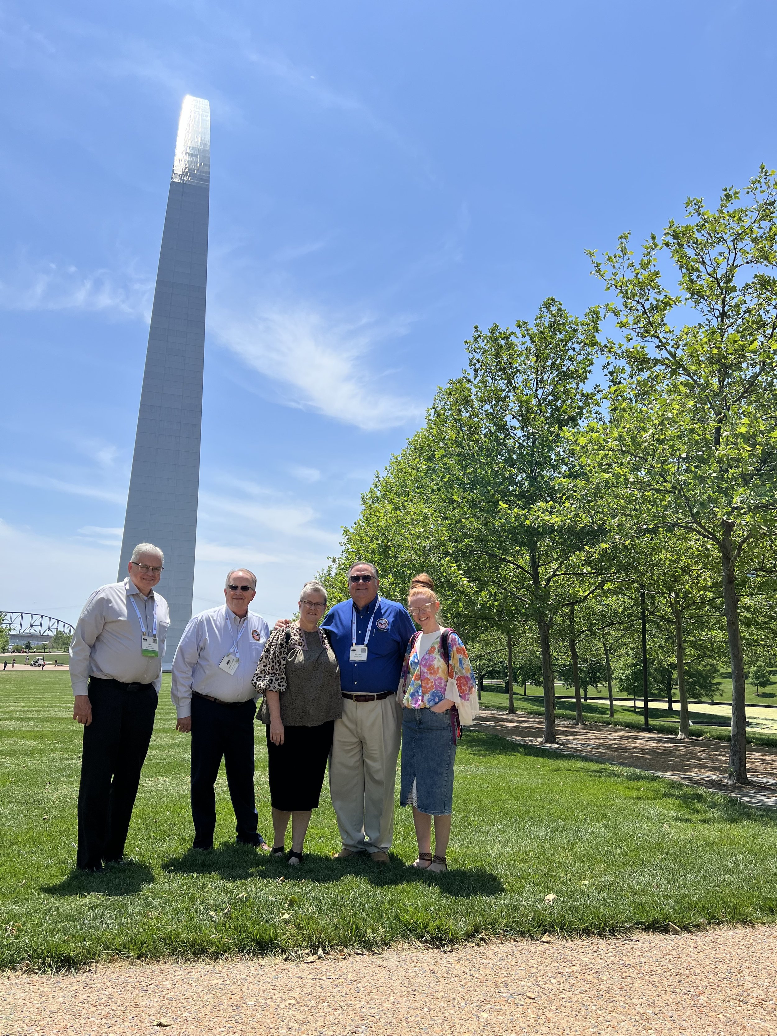 Group of six people standing outdoors on grass with green trees, a blue sky, and a tall building in the background.