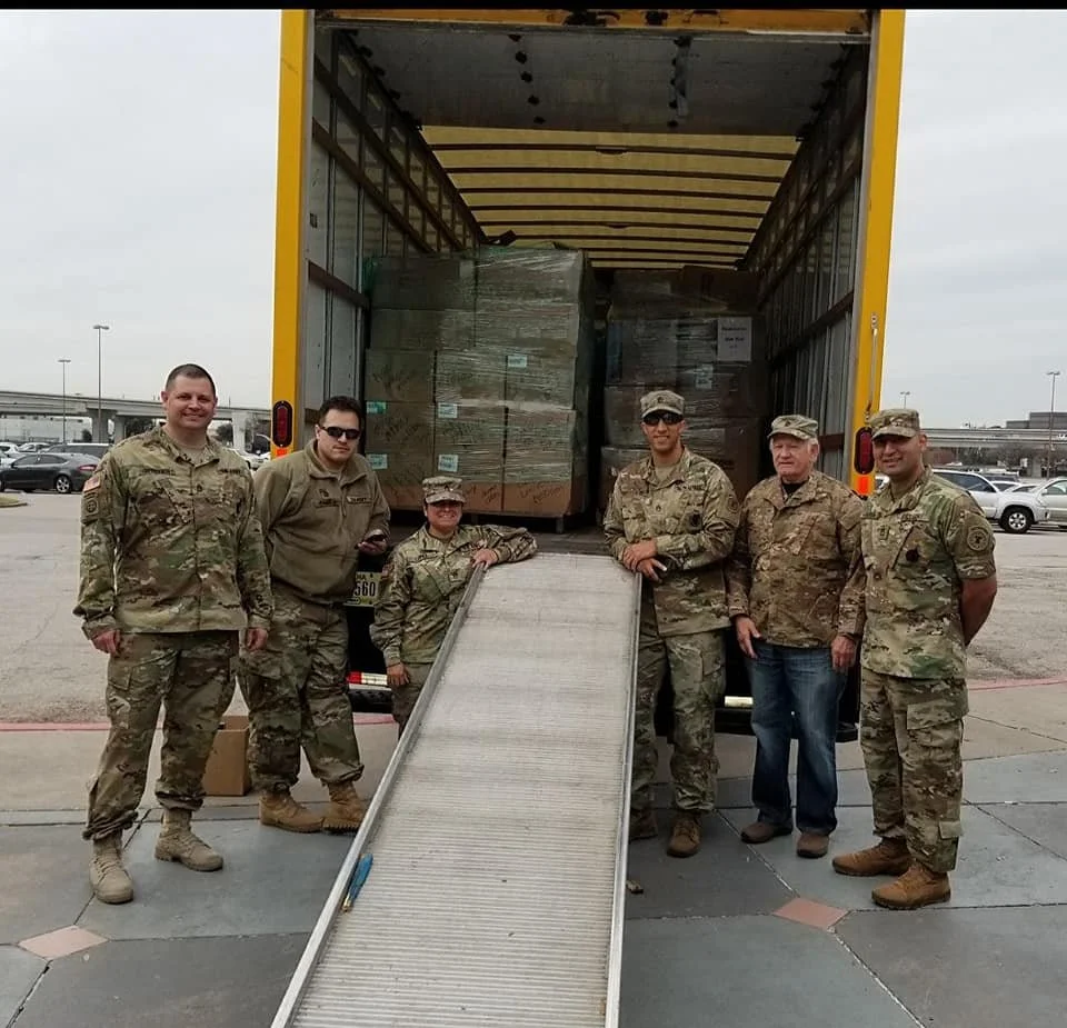 Group of seven military personnel standing in front of a truck loaded with boxes, some leaning on a ramp.