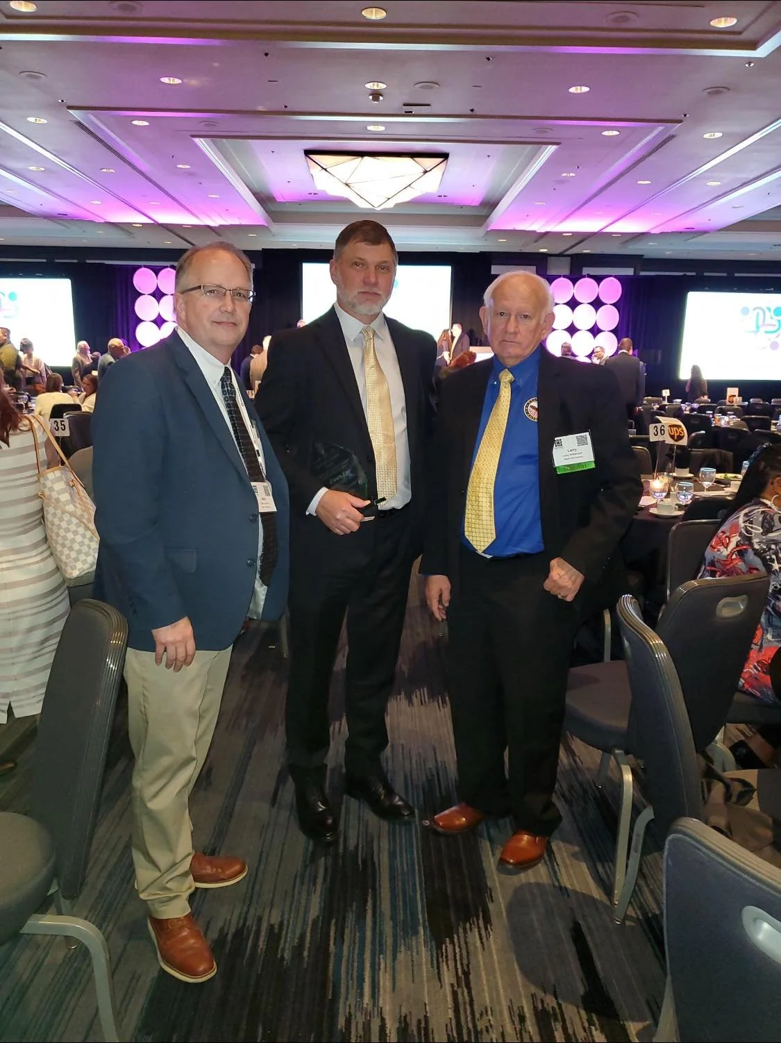 Three men in formal attire standing together at a conference or event with tables, chairs, and large screens in the background.