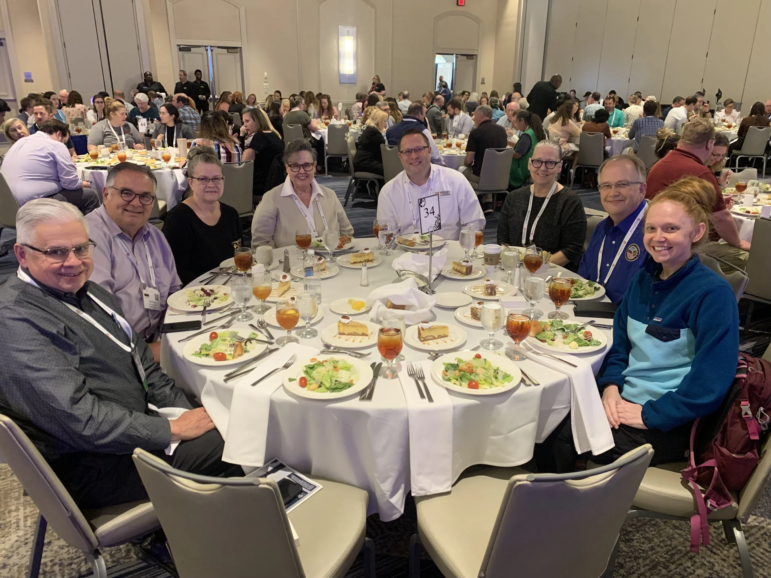 People seated around a round dining table with salads, desserts, and drinks at a banquet or conference event in a large, well-lit hall.