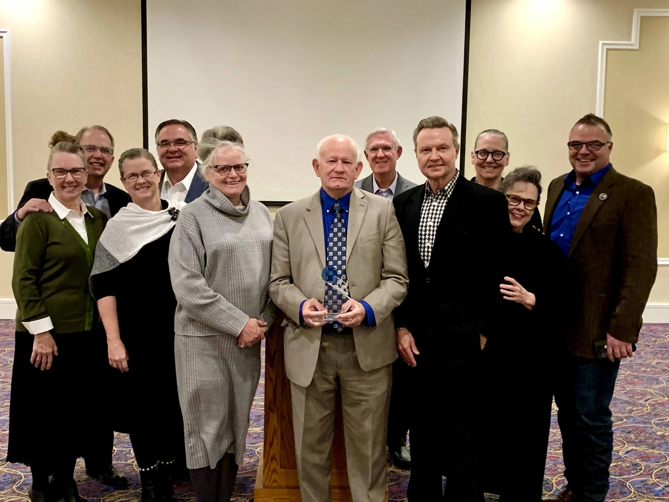 Group of twelve people posing for a photo in a conference room, with one person holding an award.