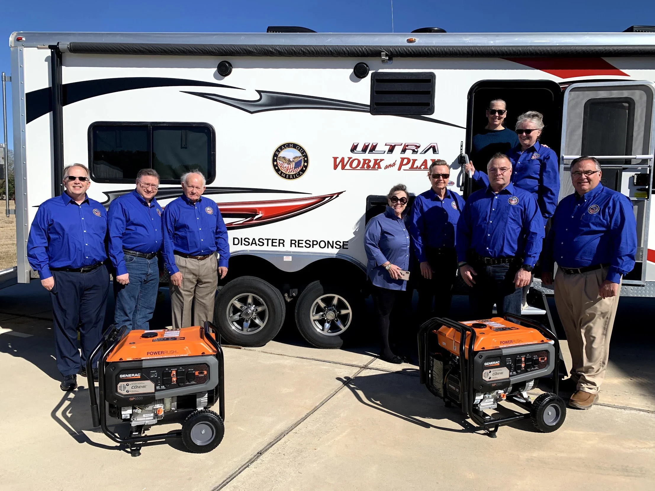 Group of ten people standing in front of a disaster response trailer with two orange portable generators on the ground.