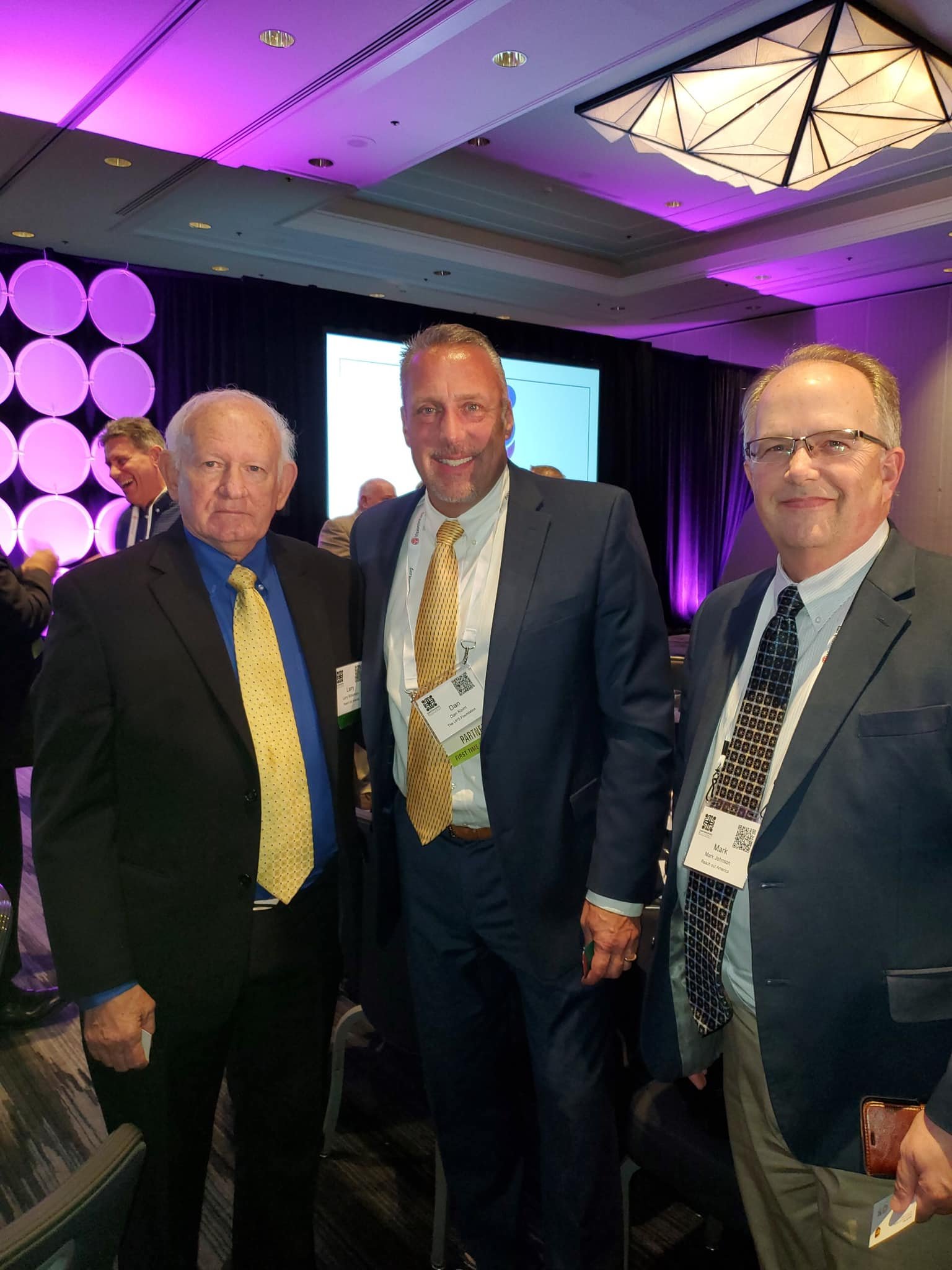 Three men in suits at a conference or event, standing together indoors with conference badges, purple lighting, and a stage in the background.