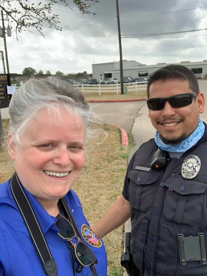 A woman photographer with sunglasses hanging from her shirt, smiling, standing next to a police officer who is wearing sunglasses, a badge, and a police uniform, outside in a parking lot area.