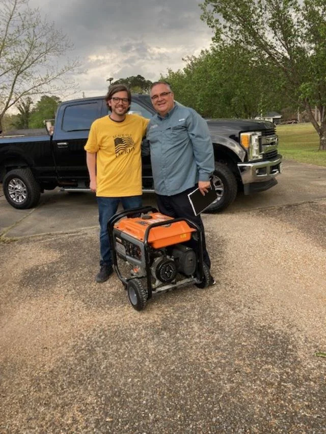 Two men standing outdoors next to a portable generator, with a black pickup truck and trees in the background under a cloudy sky.