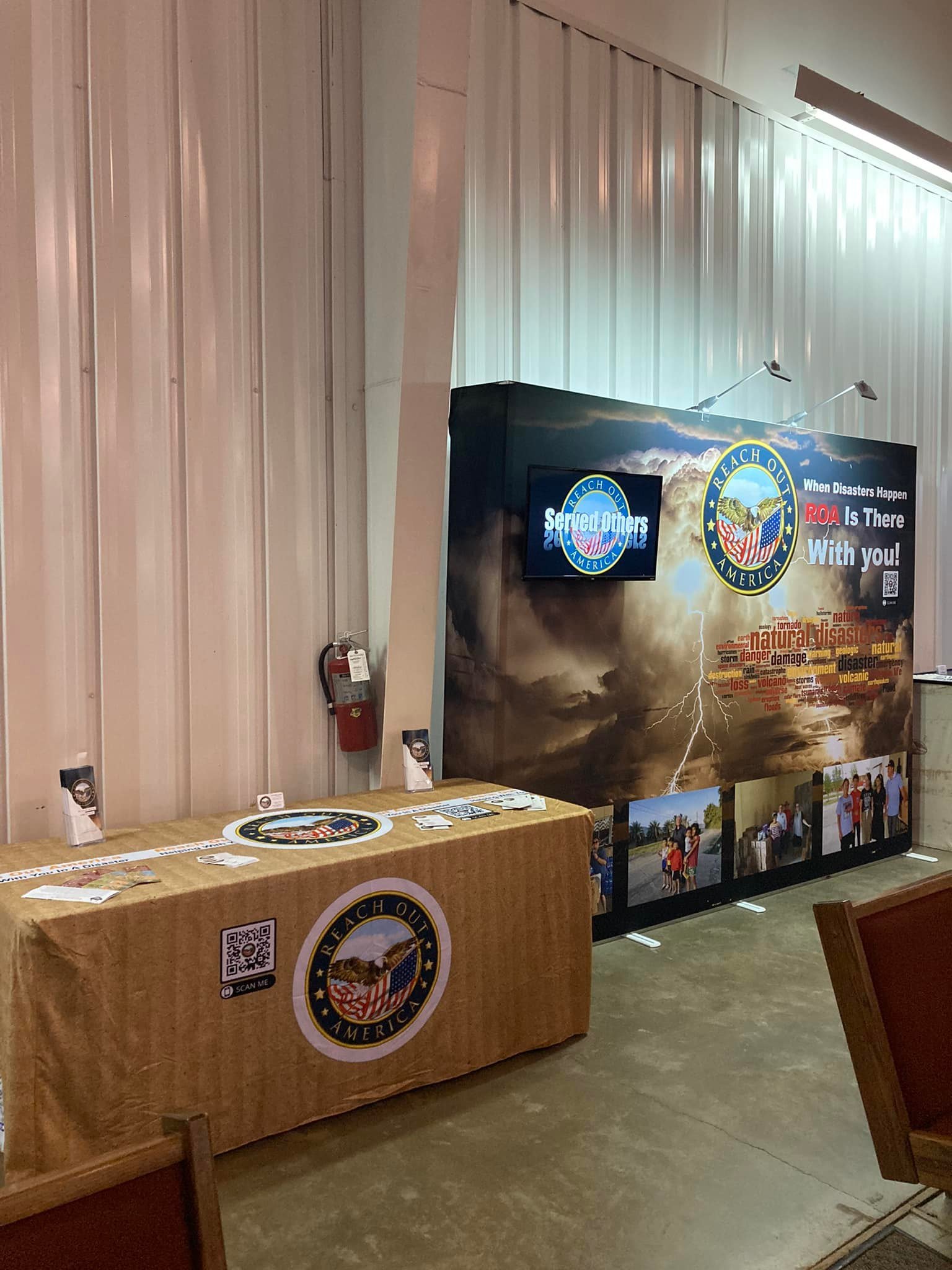 Exhibit booth with Reach Out America branding, featuring a large graphic of storm clouds and lightning, a small monitor displaying the Reach Out America logo, and photos of people in front of the exhibit.