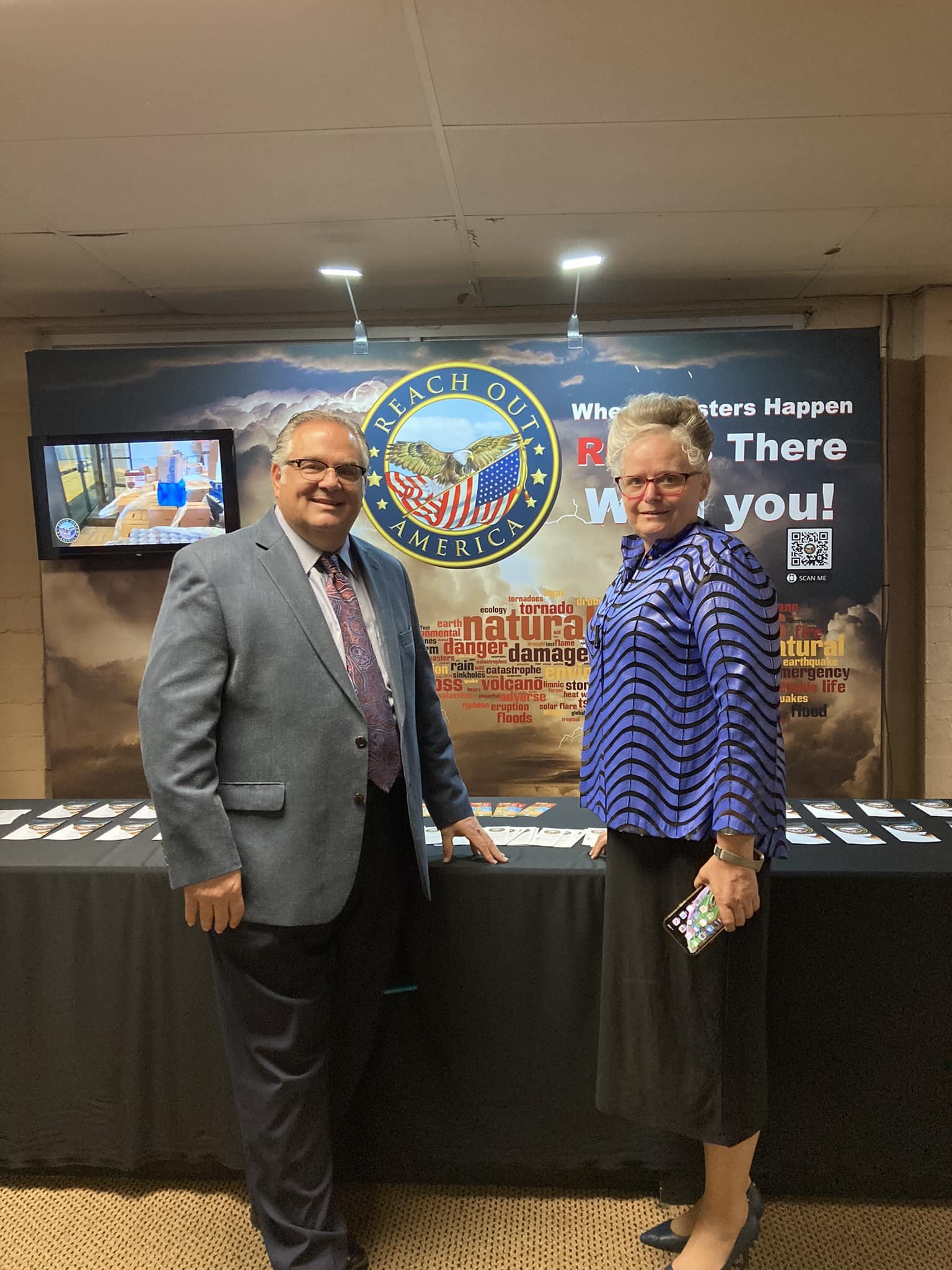 A man in a gray suit and a woman in a blue patterned jacket are standing in front of a table and a backdrop that features the REACH OUT AMERICA logo, a large eagle, and words related to natural disasters like tornado, damage, volcano, and floods. The