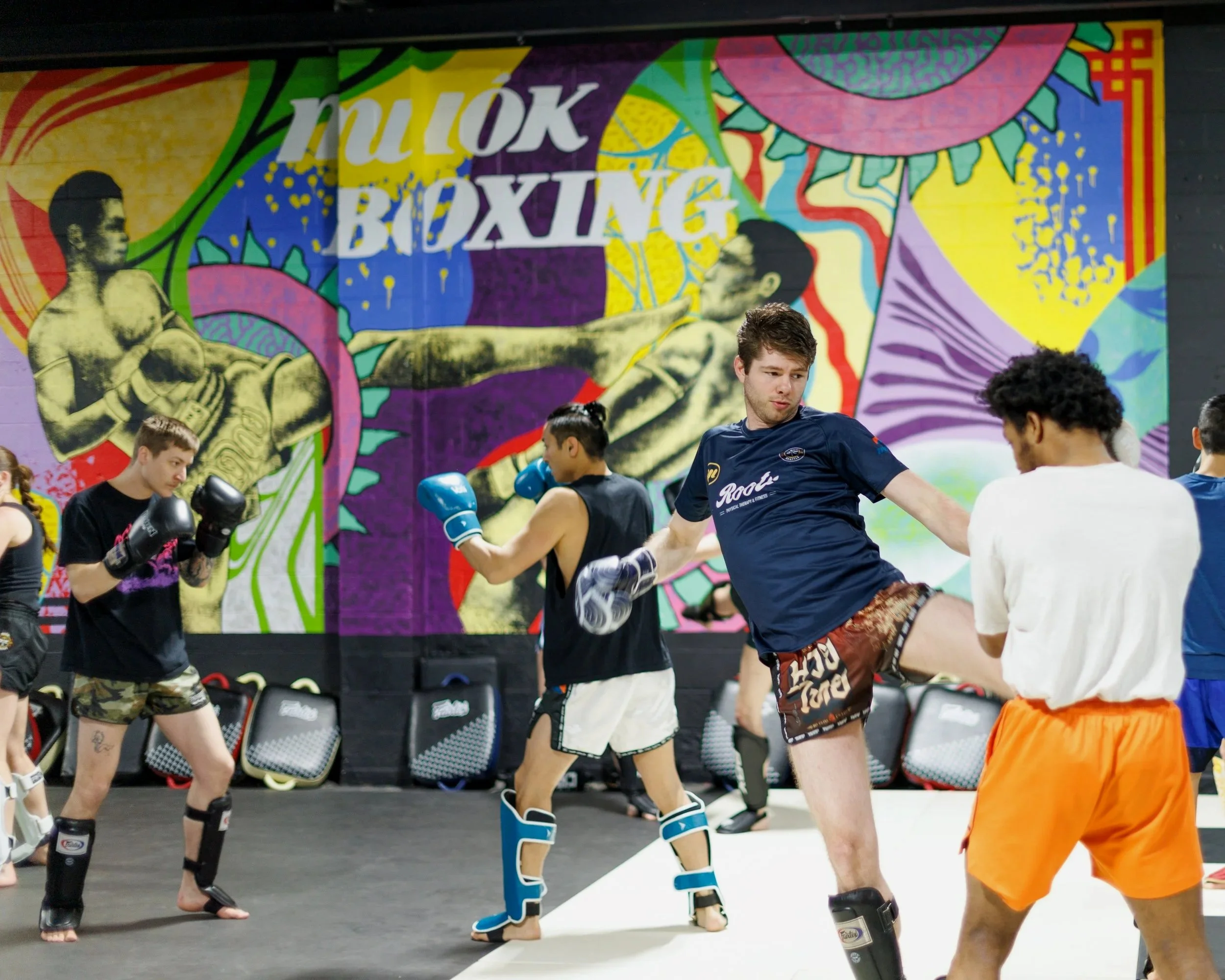 A group of people practicing kickboxing in a gym with a colorful mural of a Muay Thai boxer on the wall.