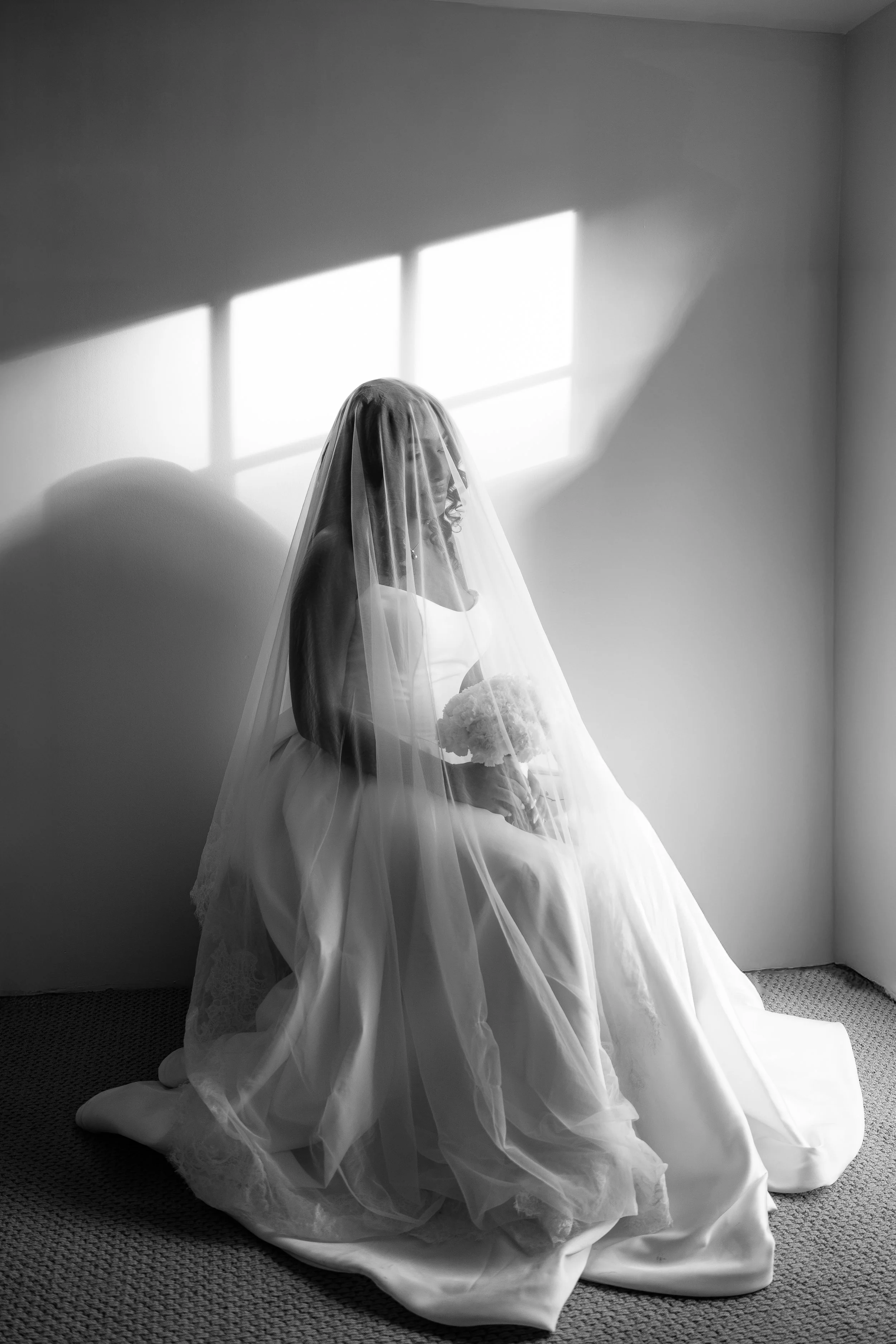 Black-and-white photo of a bride sitting on the floor in a wedding dress with a veil, holding a bouquet, with sunlight streaming through a window casting shadows on the wall in Orlando, Florida.