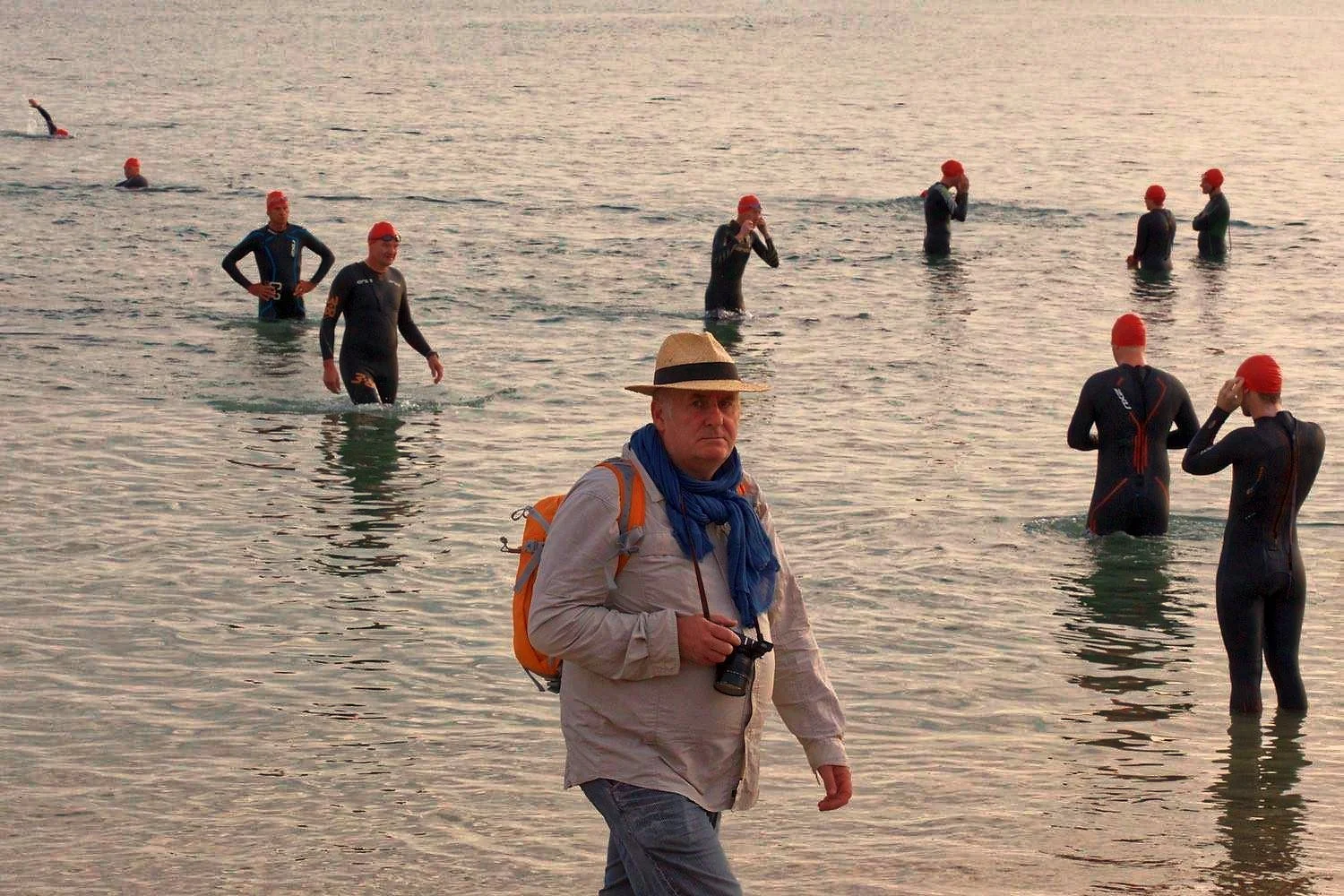 A man with a hat, beige shirt, and blue scarf standing on the beach, holding a camera, with nine swimmers in wetsuits and red swim caps in the water behind him.