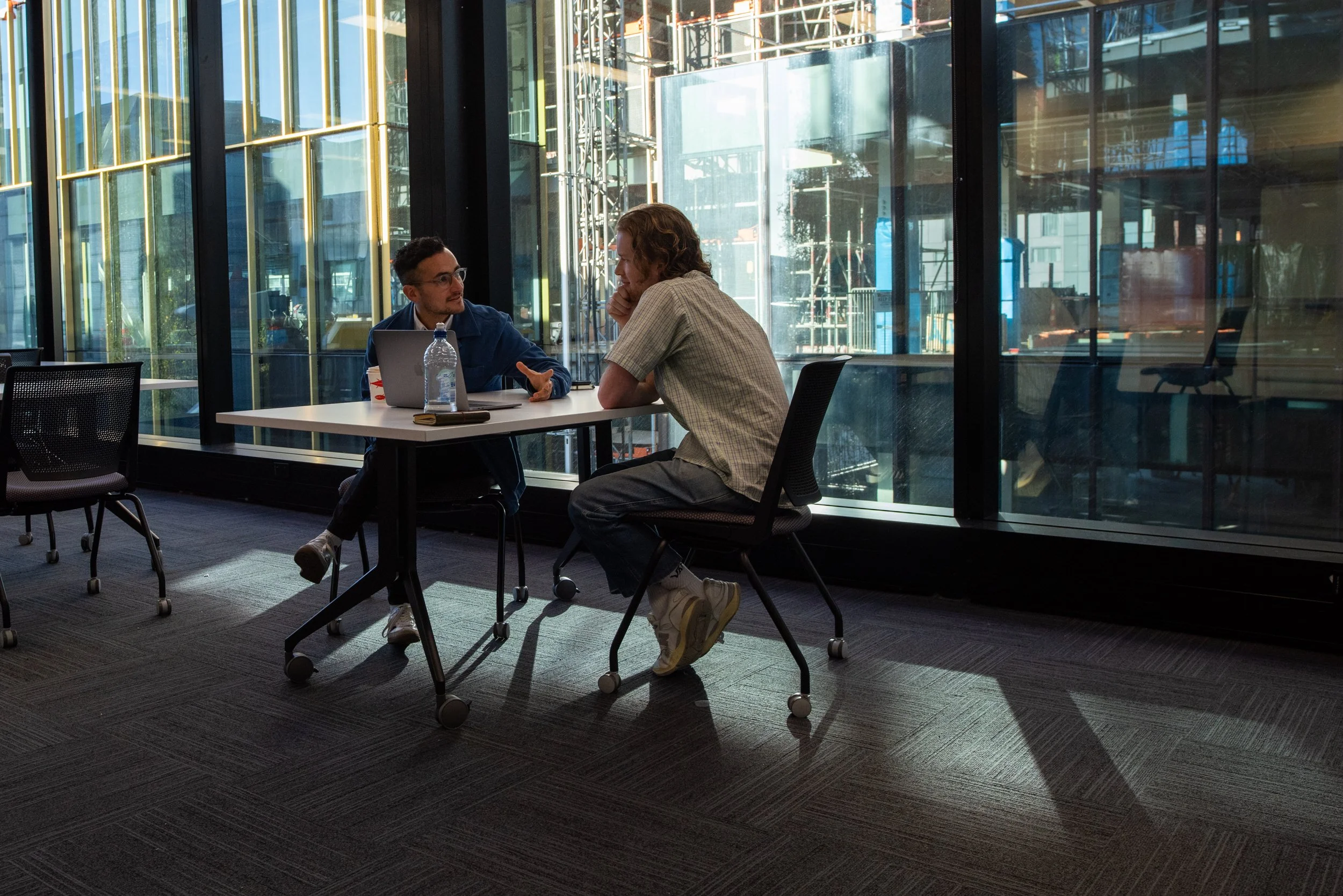 Two men having a discussion at a table in a modern office with large glass windows.