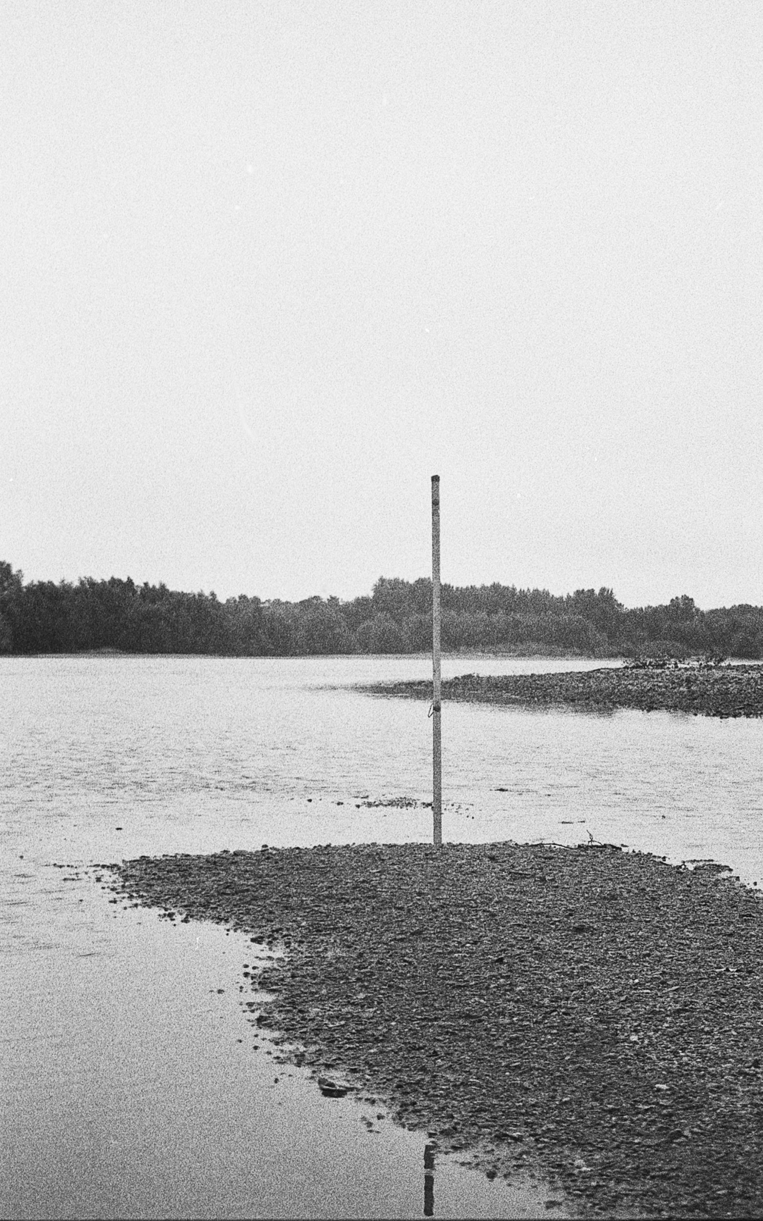 A black and white photo of a river with a makeshift flagpole on a small patch of land, surrounded by water, and trees in the distance.