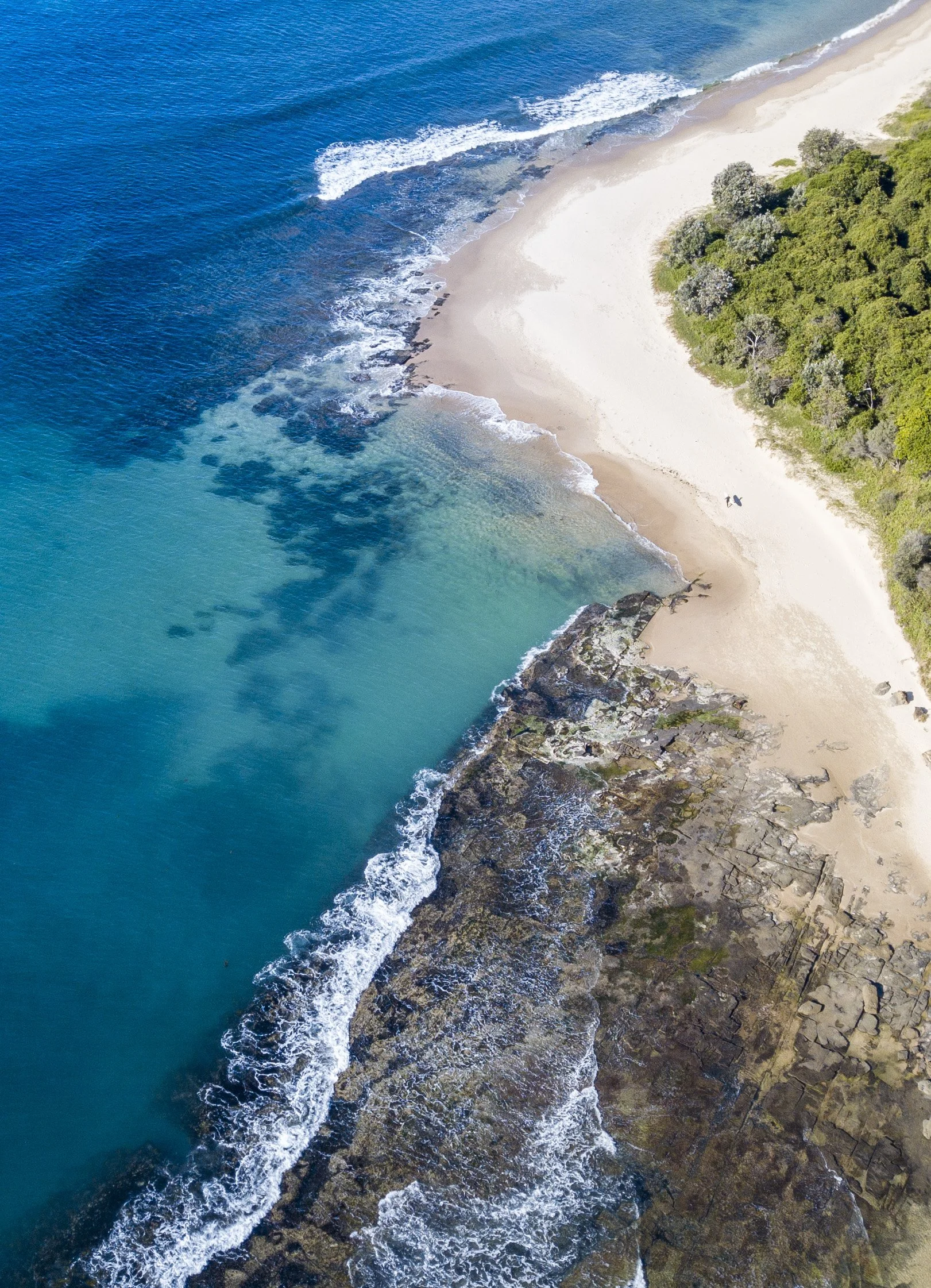 Aerial view of a beach with turquoise water, sandy shore, rocks, and green trees.