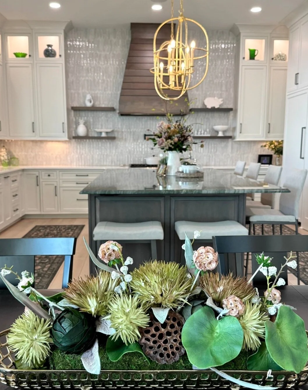 A modern, light-filled kitchen with white cabinetry, a large island with a gray base and marble top, a floral centerpiece, and a decorative chandelier.