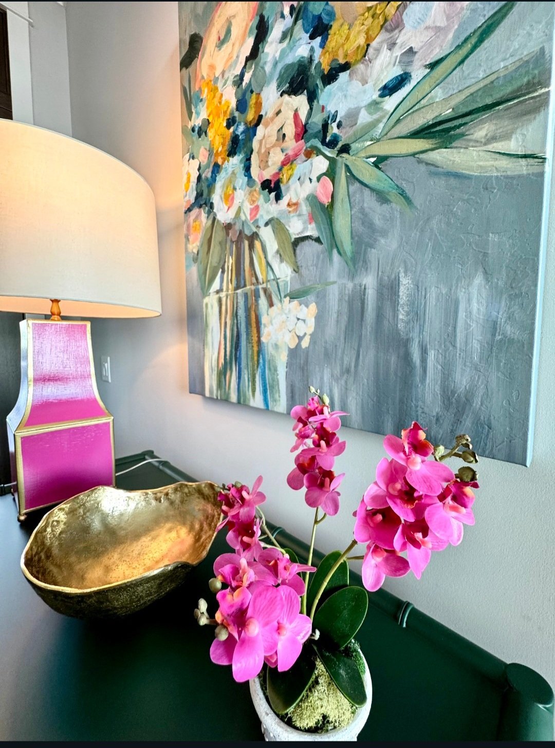 A pink orchid plant on a black table, a large beige lampshade, a colorful floral painting on the wall, and a decorative gold bowl on the table.