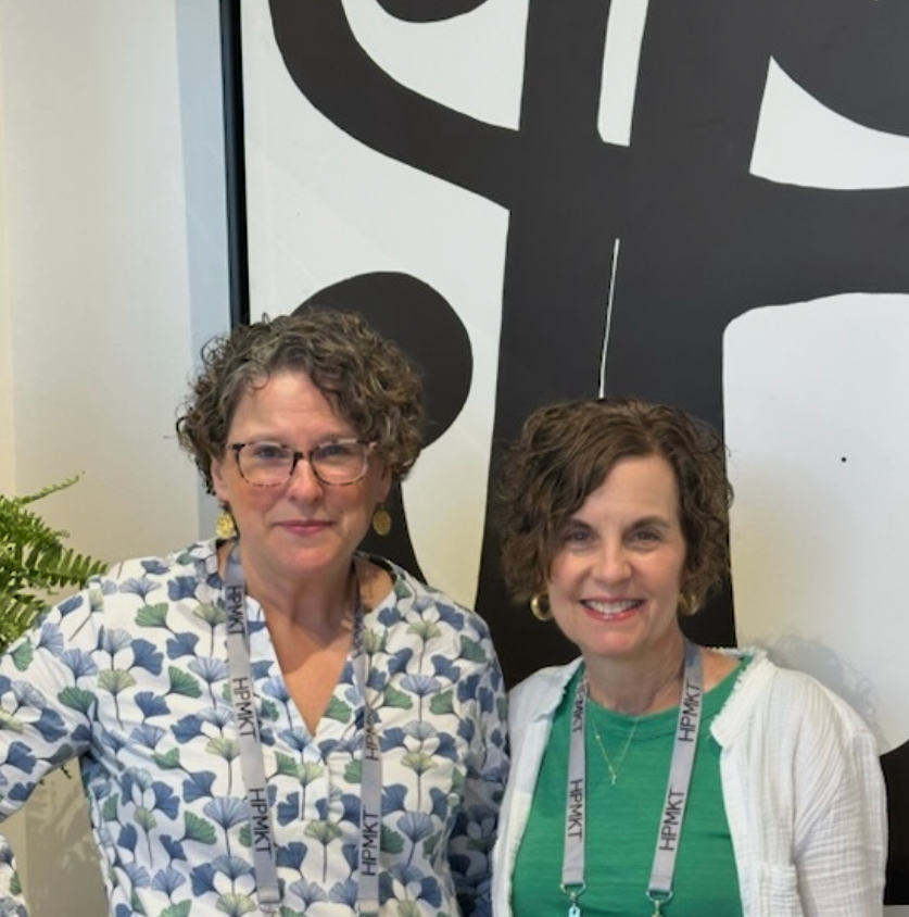 Two women with curly hair and glasses standing side by side, smiling, wearing conference tags around their necks, in front of a black and white abstract art piece.