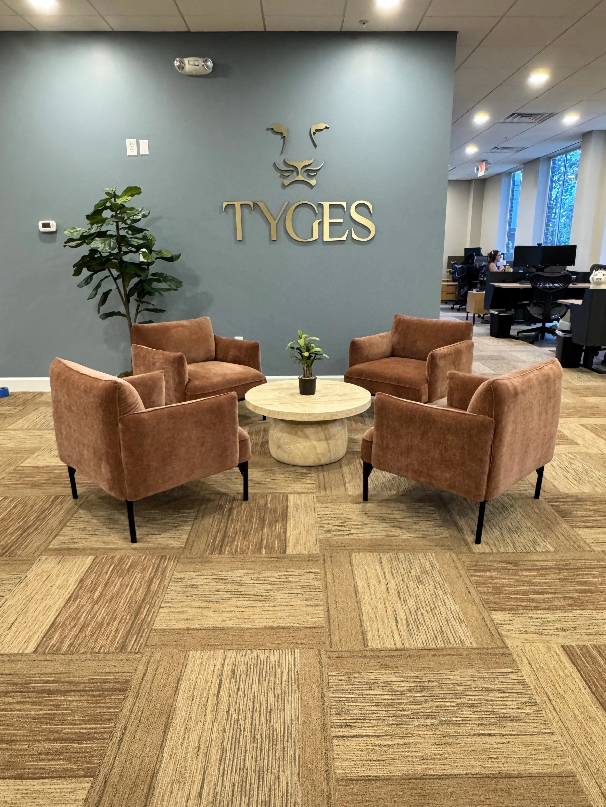 An office lounge area with four pink armchairs arranged around a small round wooden table with a small potted plant. A large green plant is next to the wall. The wall has a sign with a lion face made of metal and the word 'TYGES'. In the background, there are workstations with people working.