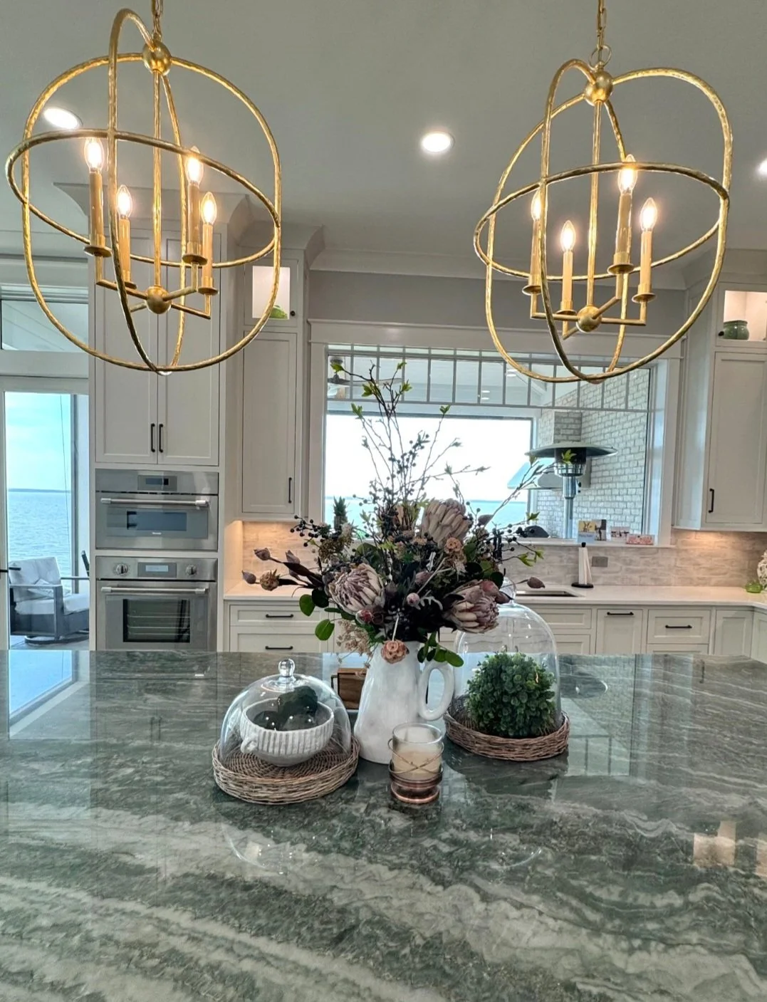 Kitchen with two gold chandeliers, floral arrangement on a green marble island, and a view of the ocean through large windows.