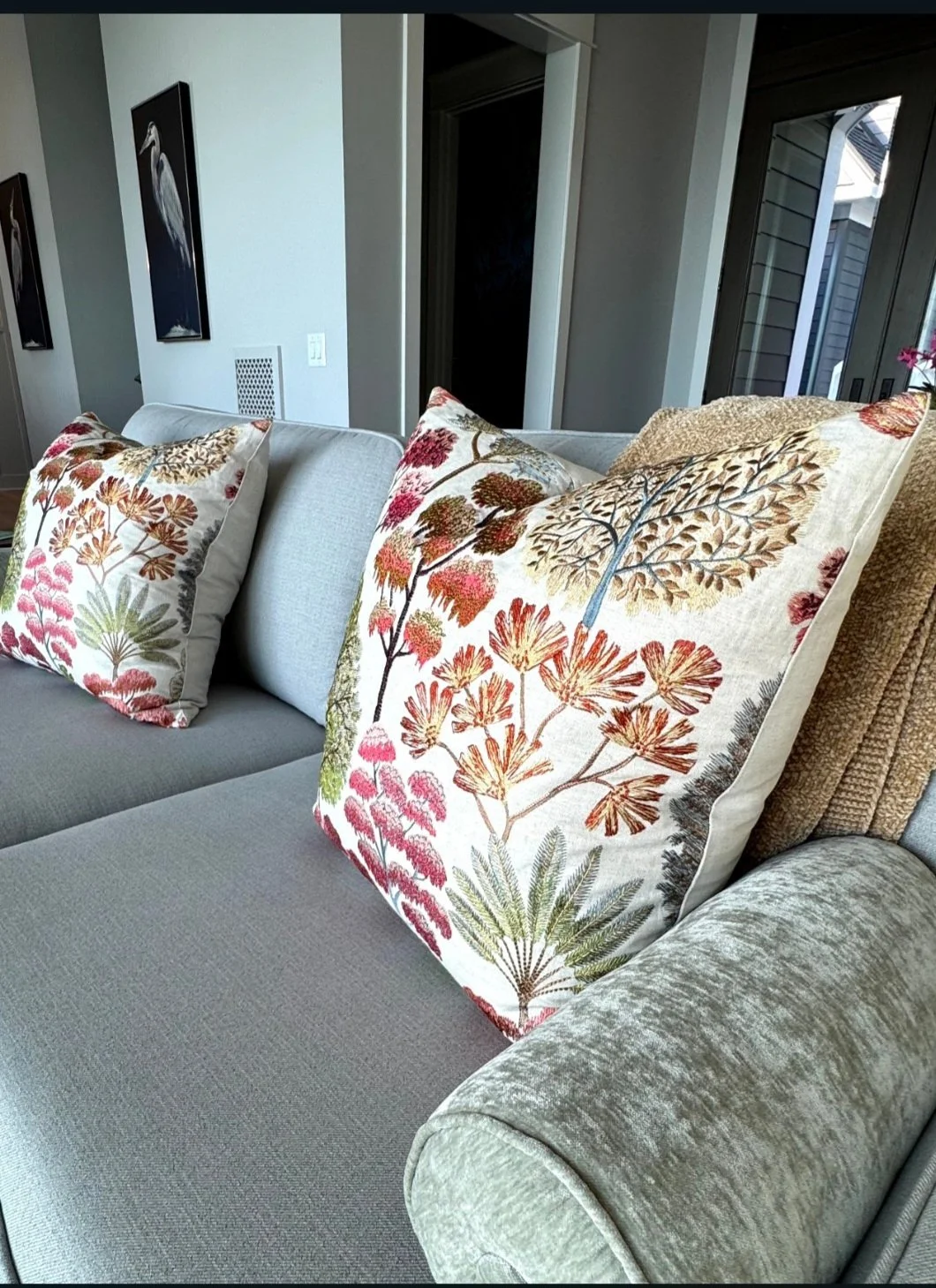 Two decorative cushions with botanical and floral embroidery on a light-colored sofa in a modern living room, with wall art and sliding glass doors in the background.