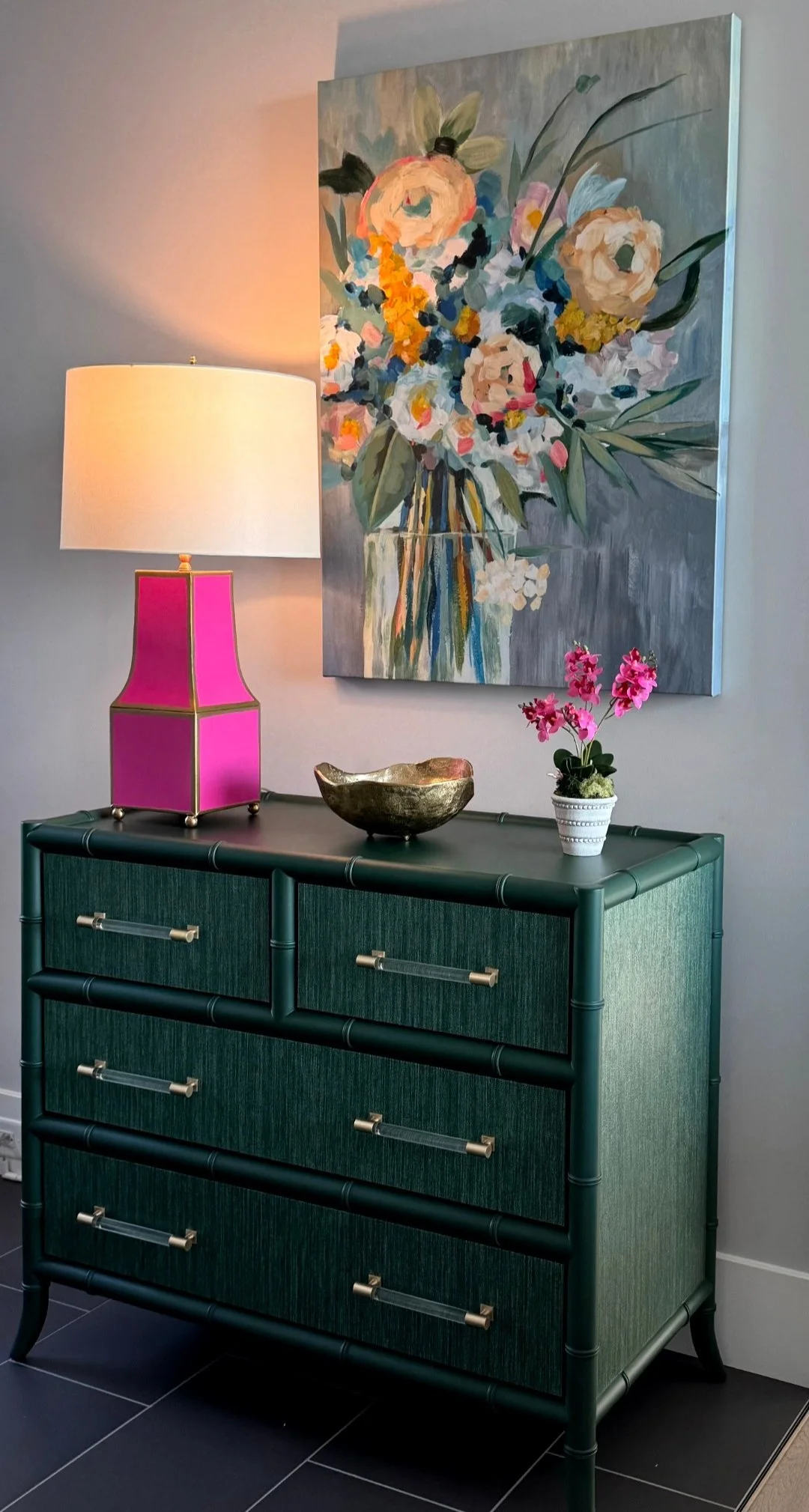 A green dresser with four drawers, decorated with a pink lamp, a decorative bowl, and a pink orchid plant, with a large floral painting on the wall behind.