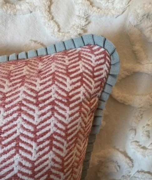 Close-up of a pillow with a pink and white chevron pattern, bordered by blue trim, against a textured off-white wall.