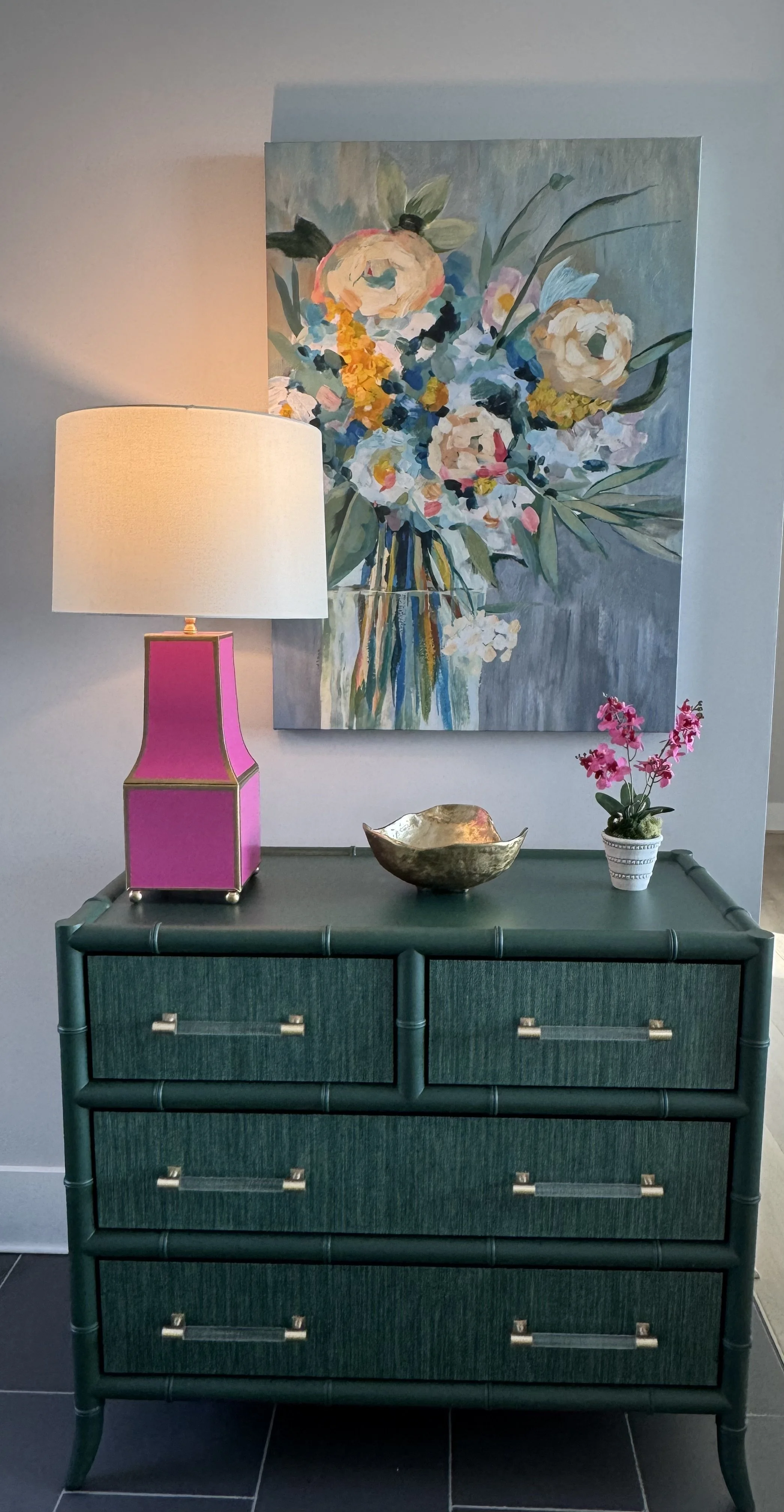 Decorative teal dresser with silver handles, topped with a pink lamp, a metallic bowl, and a small pink flower plant in a white pot; behind a large floral painting on the wall.