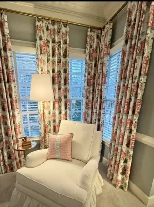 A cozy corner with floral curtains, a white armchair with a cushion, a table holding a wooden tray, and a tall floor lamp in a well-lit room.