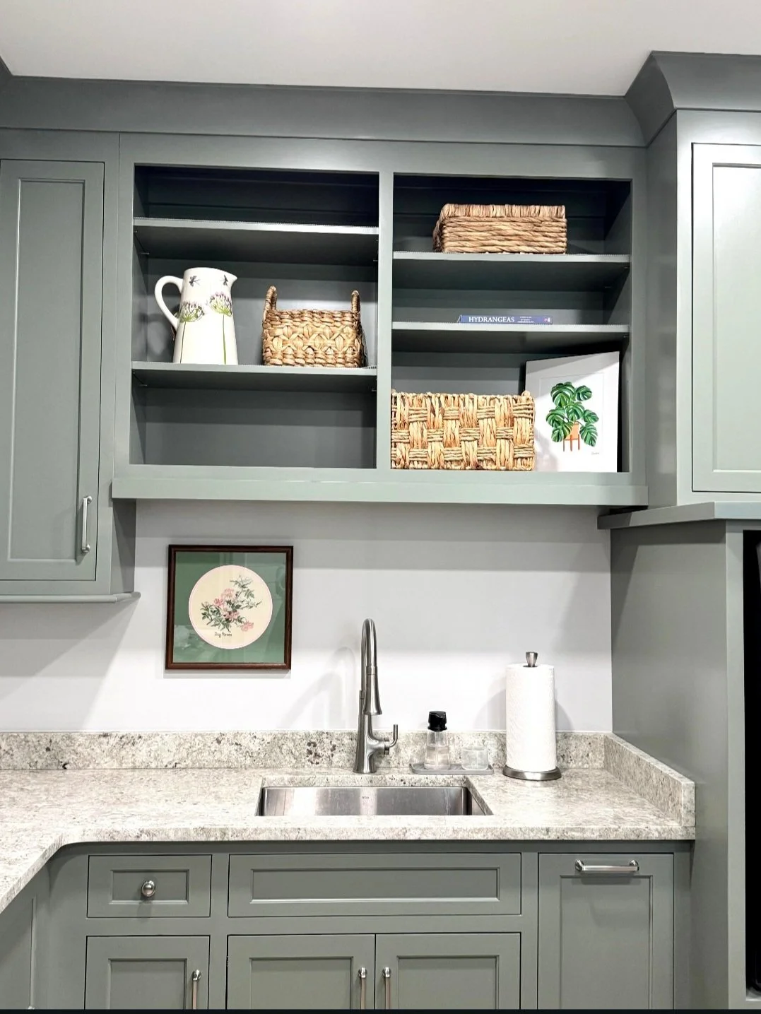 Kitchen with gray cabinets, open shelves, a granite countertop, a sink with a brushed nickel faucet, a framed botanical art piece on the wall, and decorative baskets and books on the shelves.