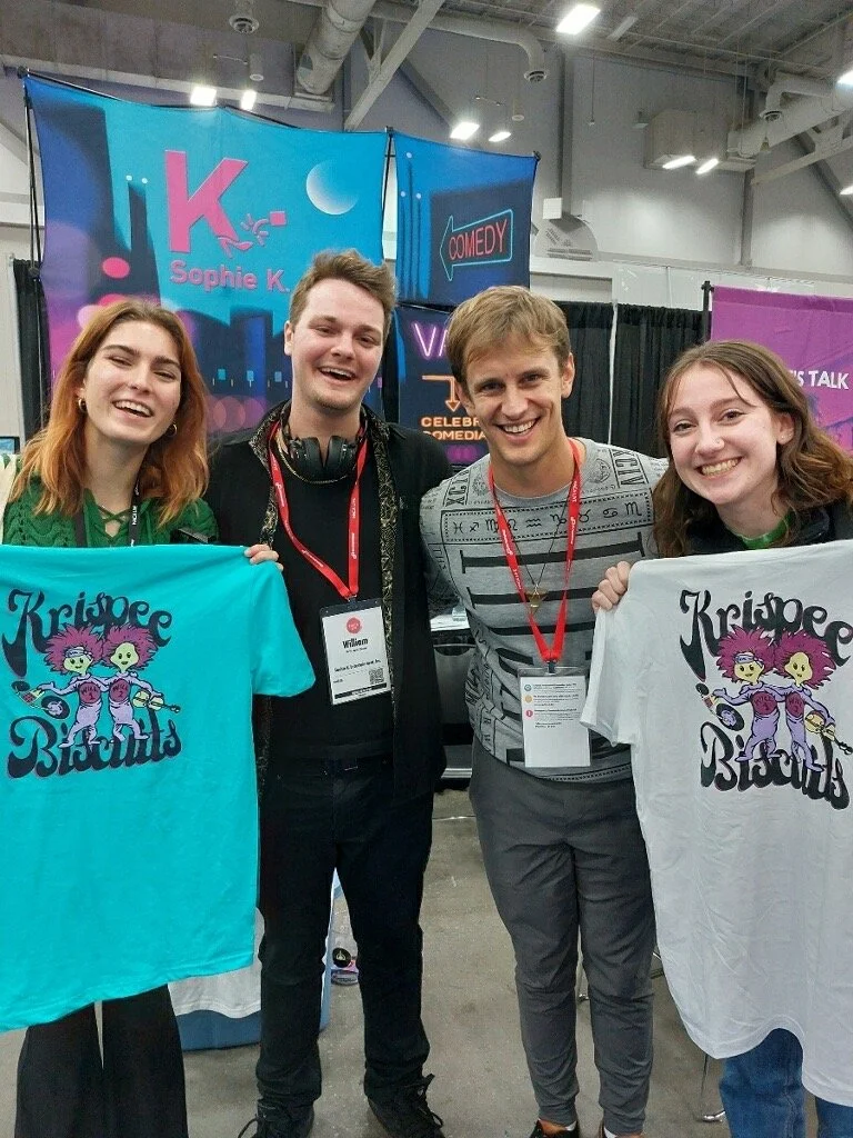 Four people at a convention holding Krispee Biscuits t-shirts in front of a booth. Fans. Event. Music. Fun. Meet and greet. party. live music. support. merch. buy.