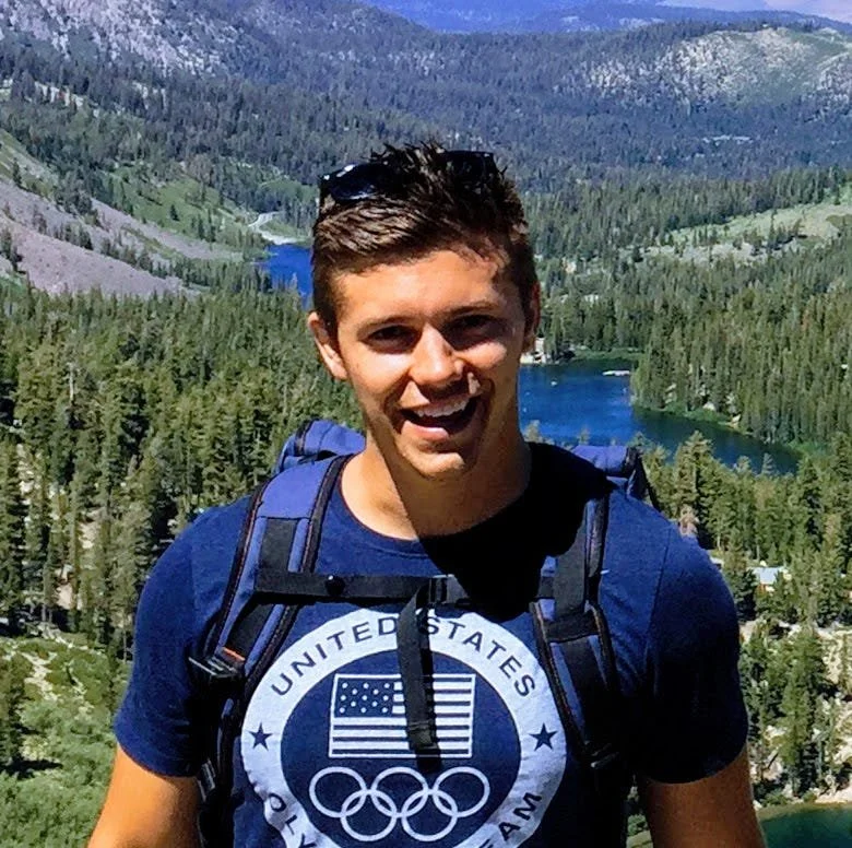 Young man smiling in front of a mountain lake with forests and hills in the background, wearing a blue United States Olympic shirt and a hiking backpack.