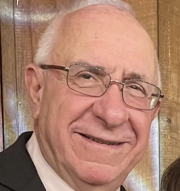 A smiling elderly man with glasses, short white hair, wearing a suit, standing in front of a wooden background.