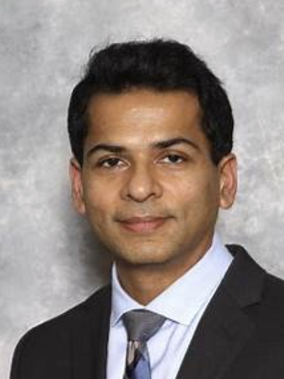 A professional headshot of a man in a black suit, white shirt, and striped tie against a gray background.