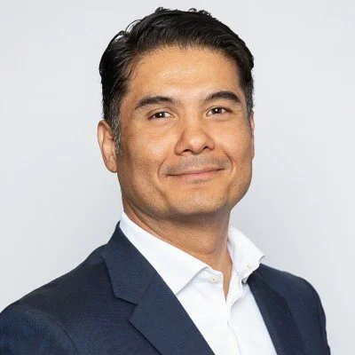 A man in a navy suit and white shirt standing against a plain light gray background.