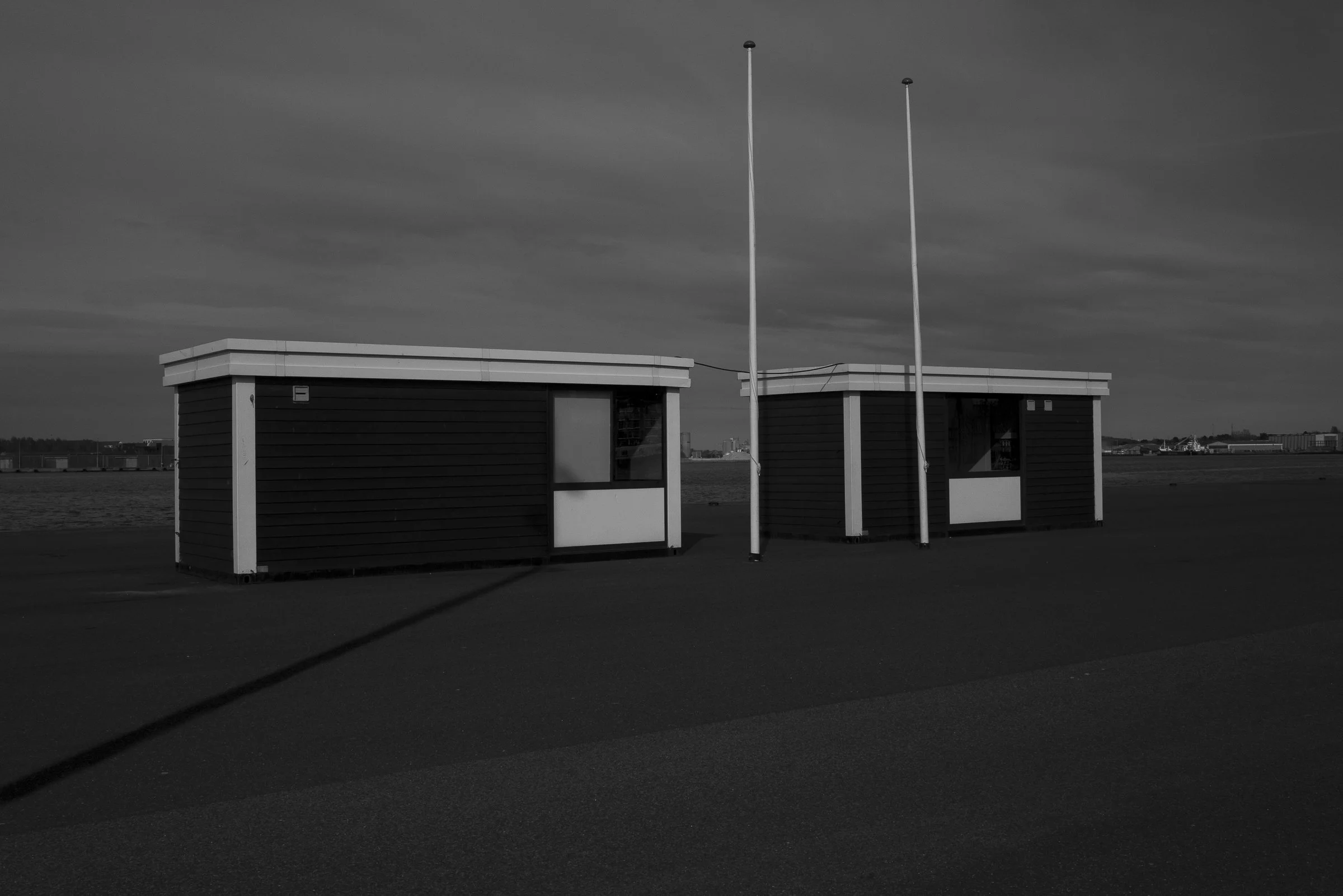 Two small beach side huts with dark siding and white trim, and two flagpoles without flags, are situated on a paved surface near the water under a cloudy sky.