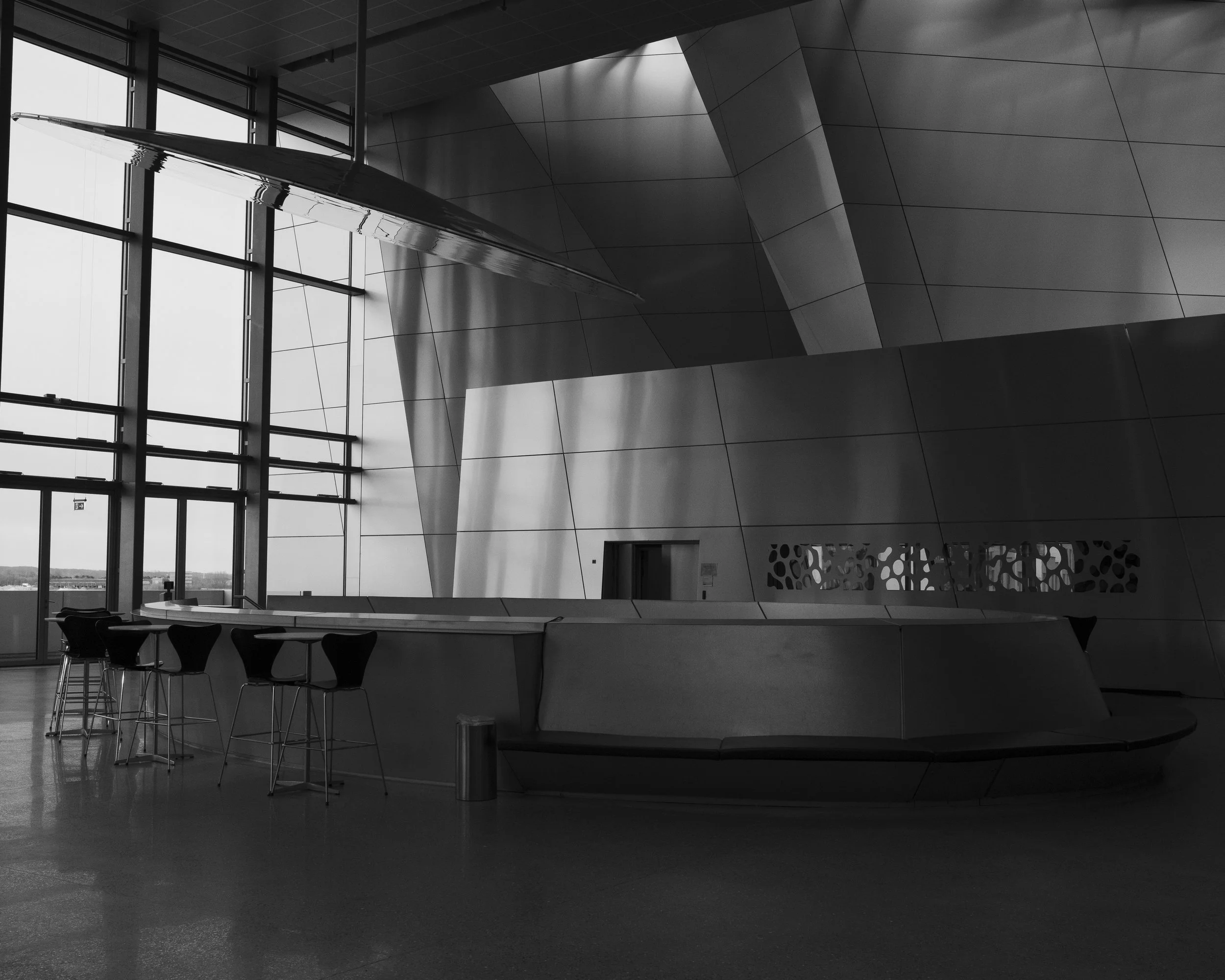Interior view of a modern building with sleek metal walls, large glass windows, a curved bar counter, and high chairs inside an architectural space.