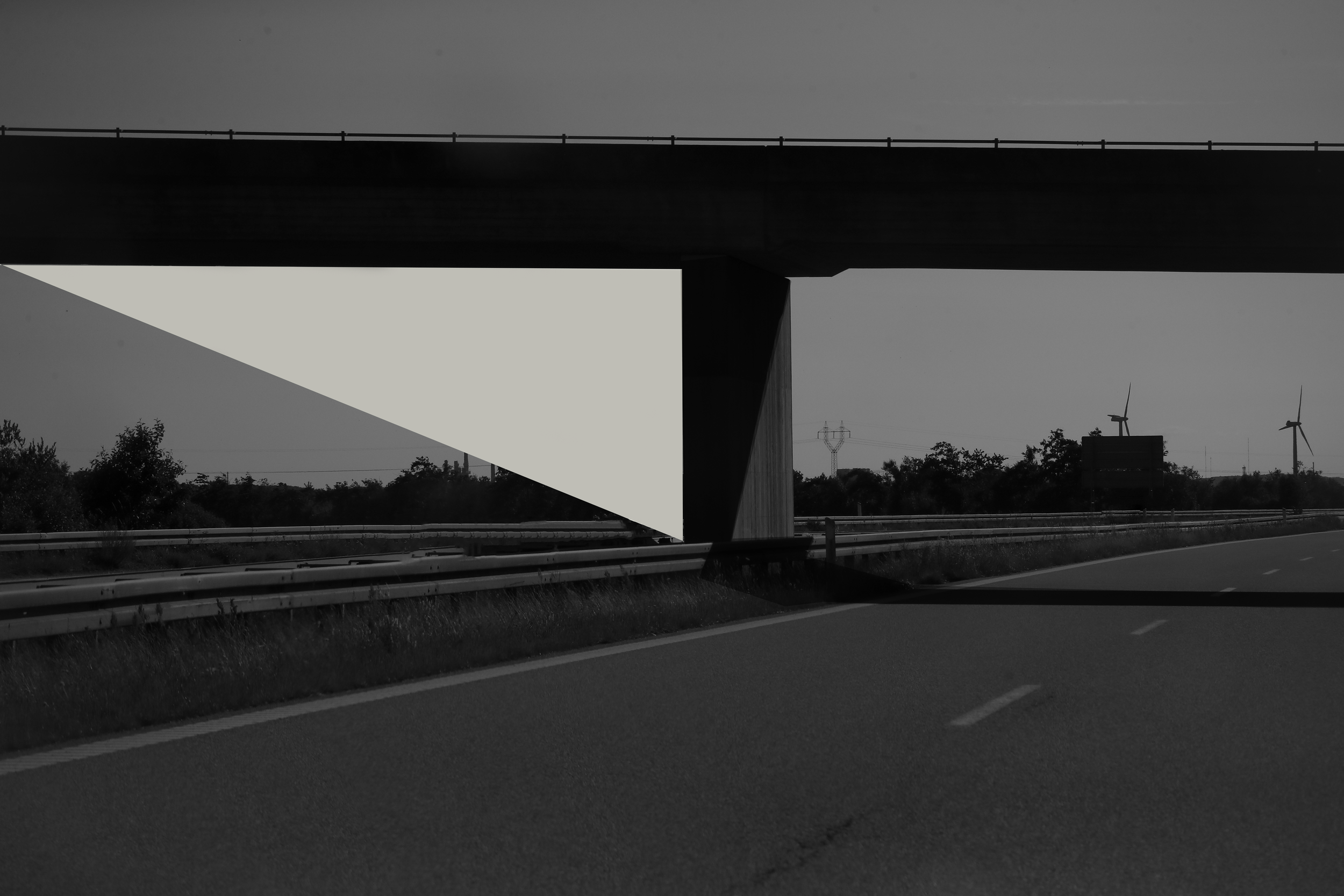 Black and white photo of an overpass bridge with a geometric white shape underneath, wind turbines in the distance, and roadside grass.