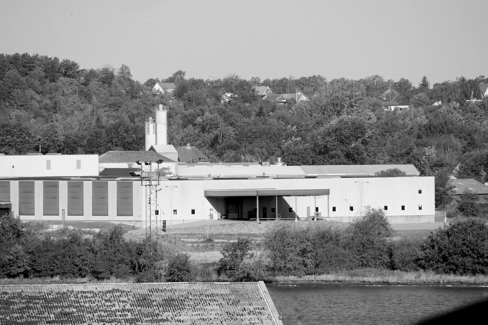 Black and white photo of an industrial building near a river, with trees and residential houses on hills in the background.