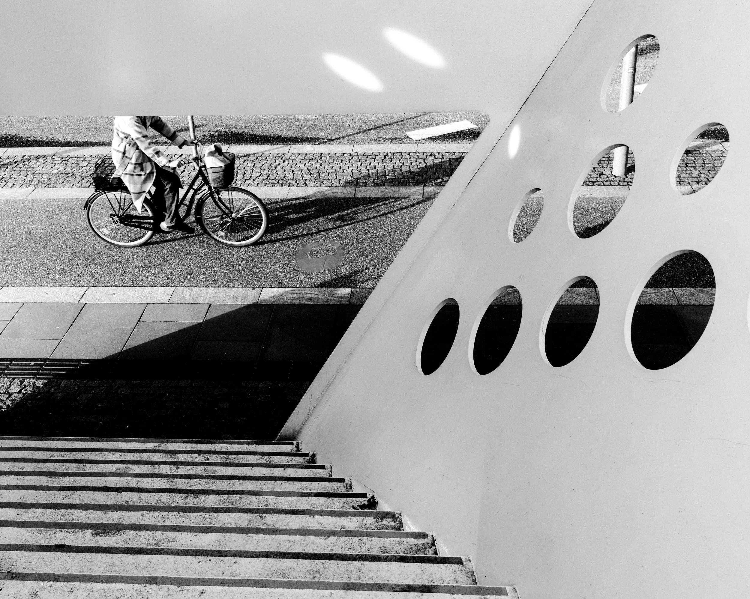 Black and white photo of a person riding a bicycle on a city street, viewed from above, with stairs and a perforated wall in the foreground.