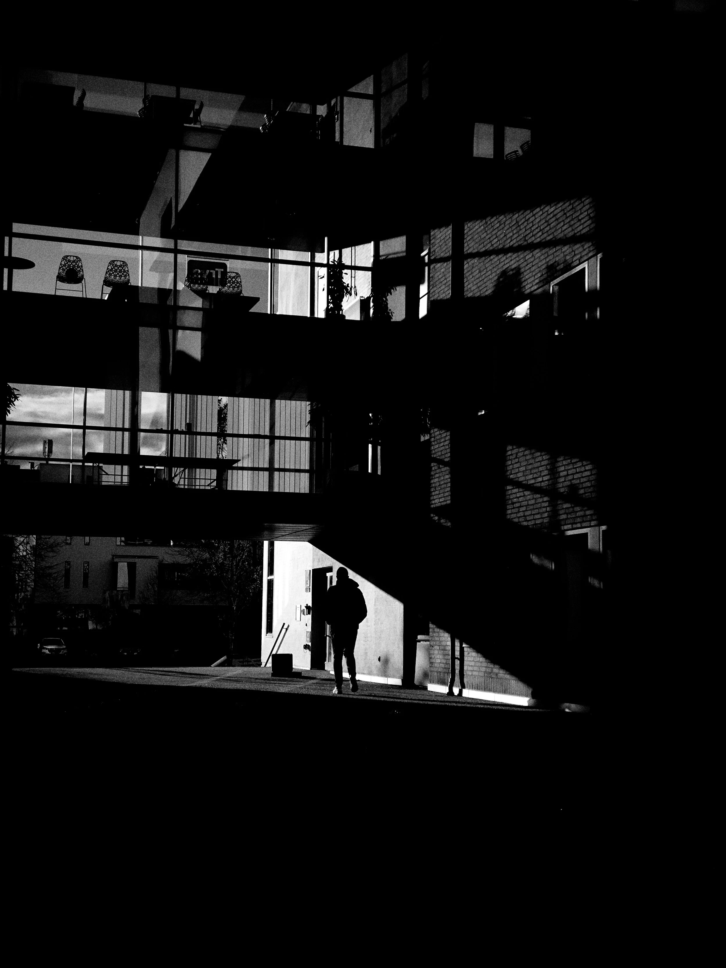 Silhouette of a person walking near a modern building with reflective glass balconies, taken in black and white with strong shadows.